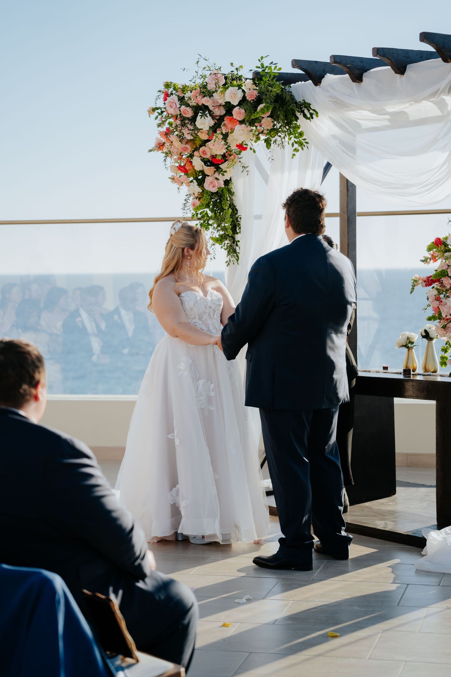 Bride and groom exchange vows under floral arch with ocean view backdrop at outdoor wedding ceremony.