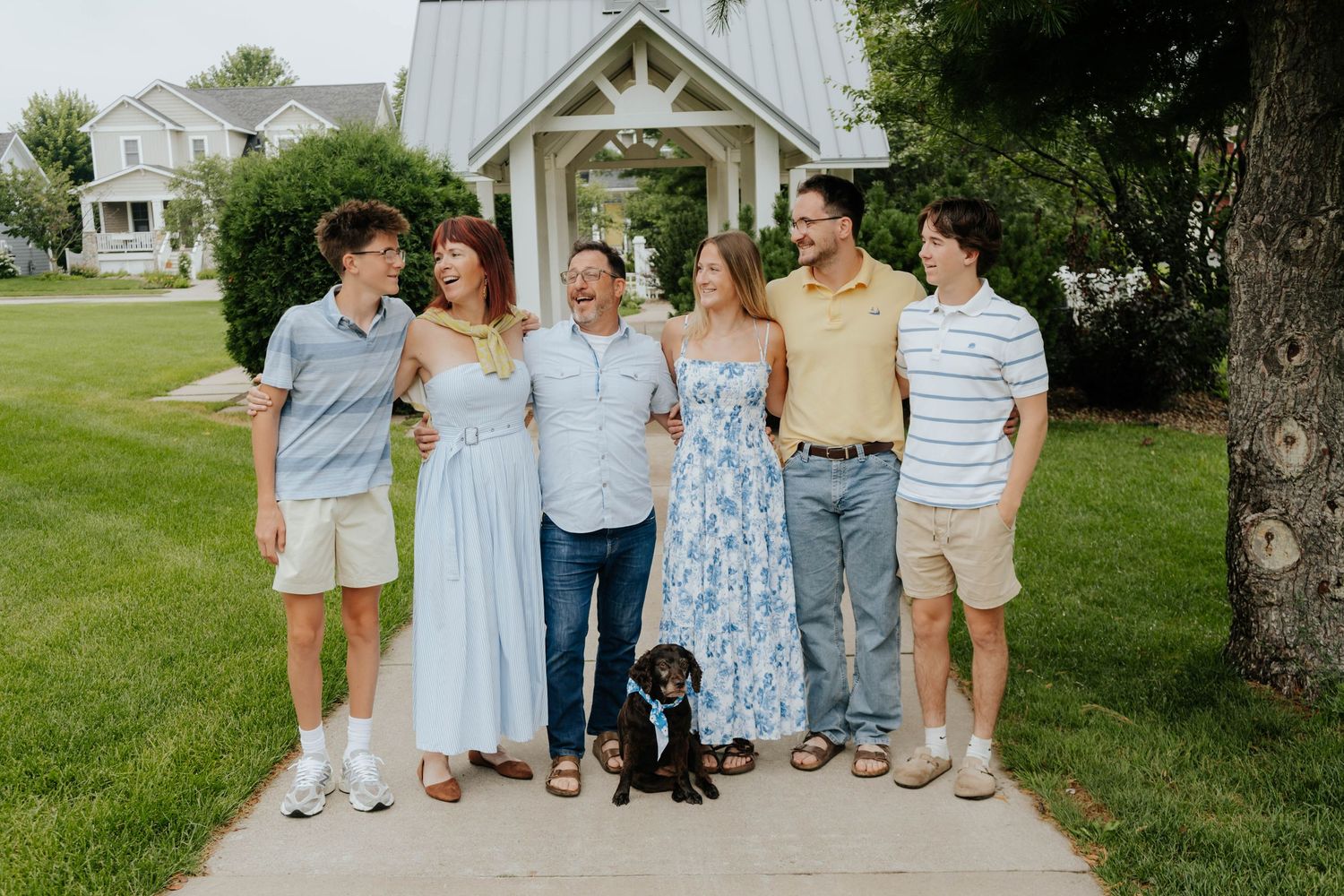 Family of six poses with small black dog on garden path near white chapel surrounded by green lawn.