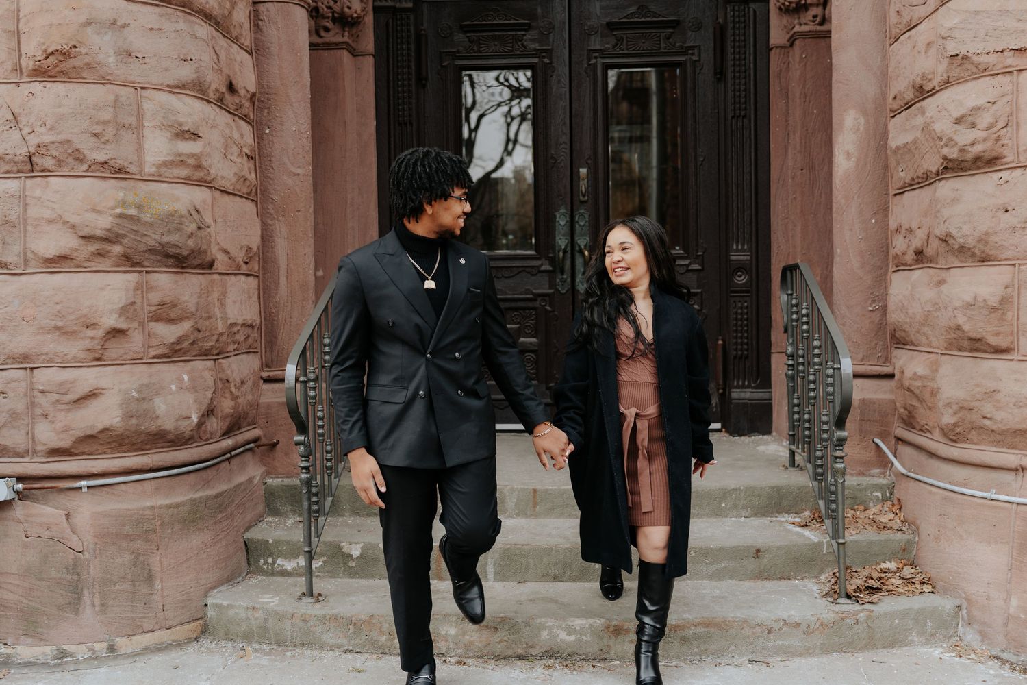 Couple holding hands and smiling while walking in front of a historic brownstone building.