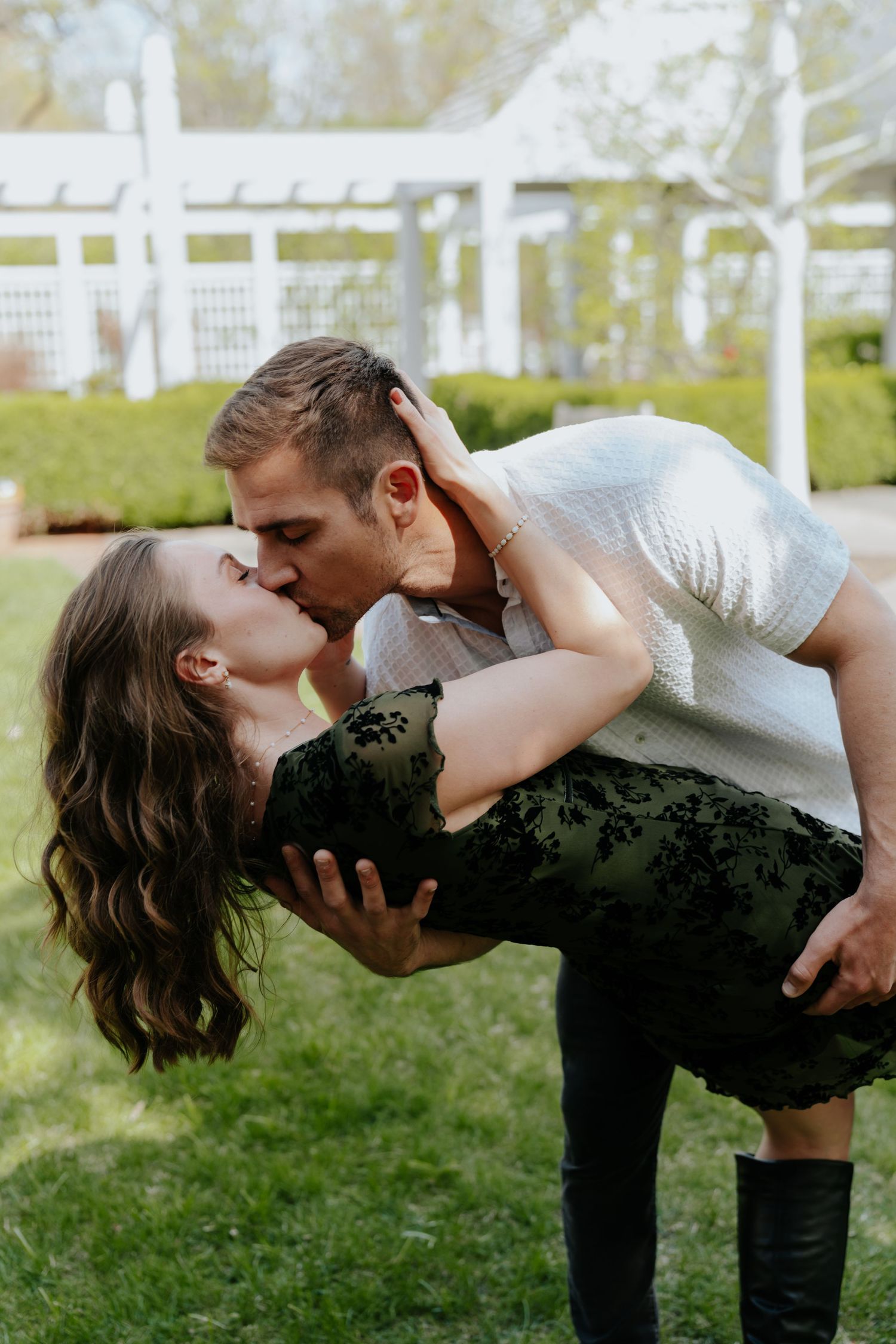 Couple sharing a passionate dip kiss outdoors on green grass with white garden fence background.