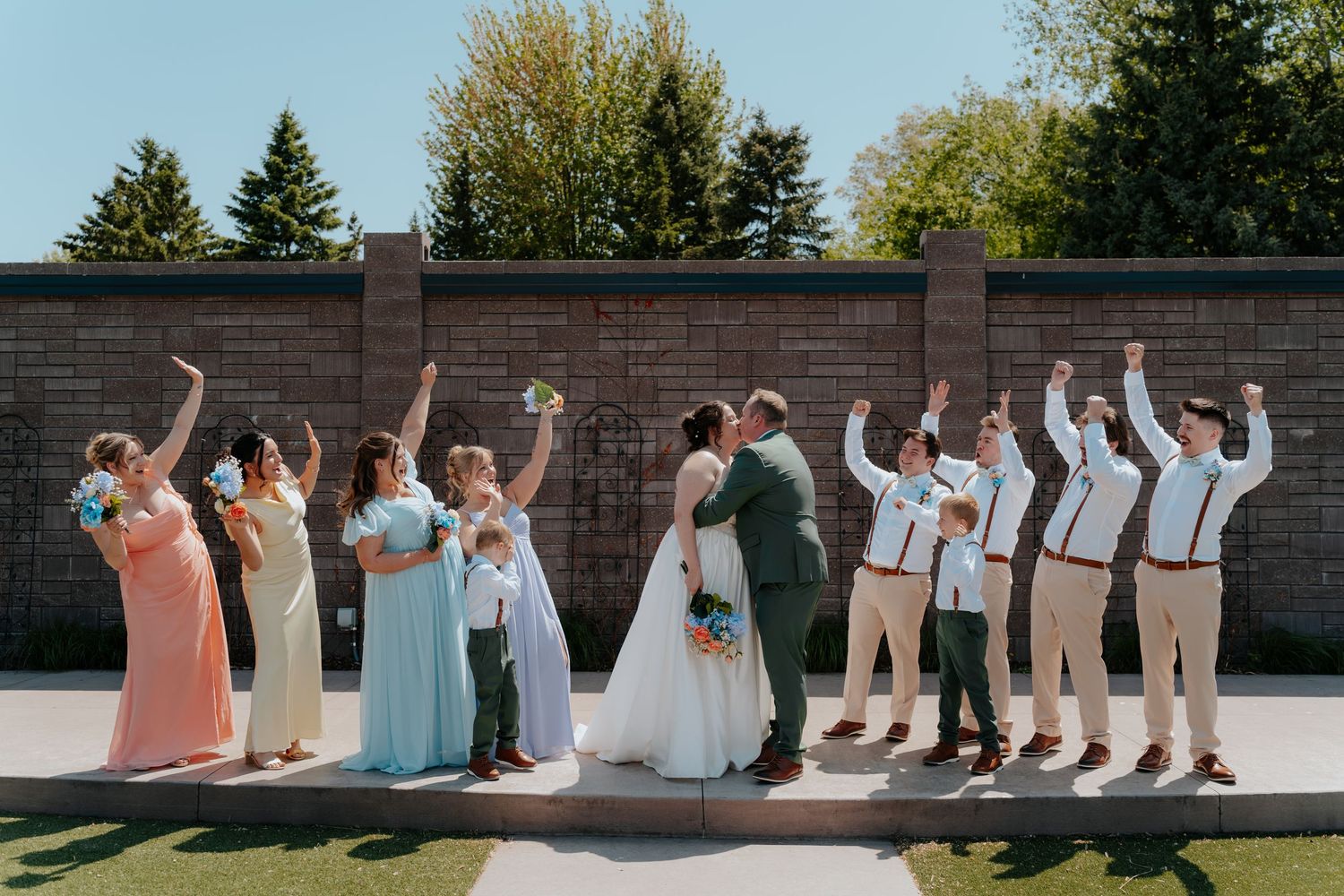 Wedding party celebrating outdoors, bridesmaids in pastel gowns and groomsmen in tan suits cheering beside bride and groom.
