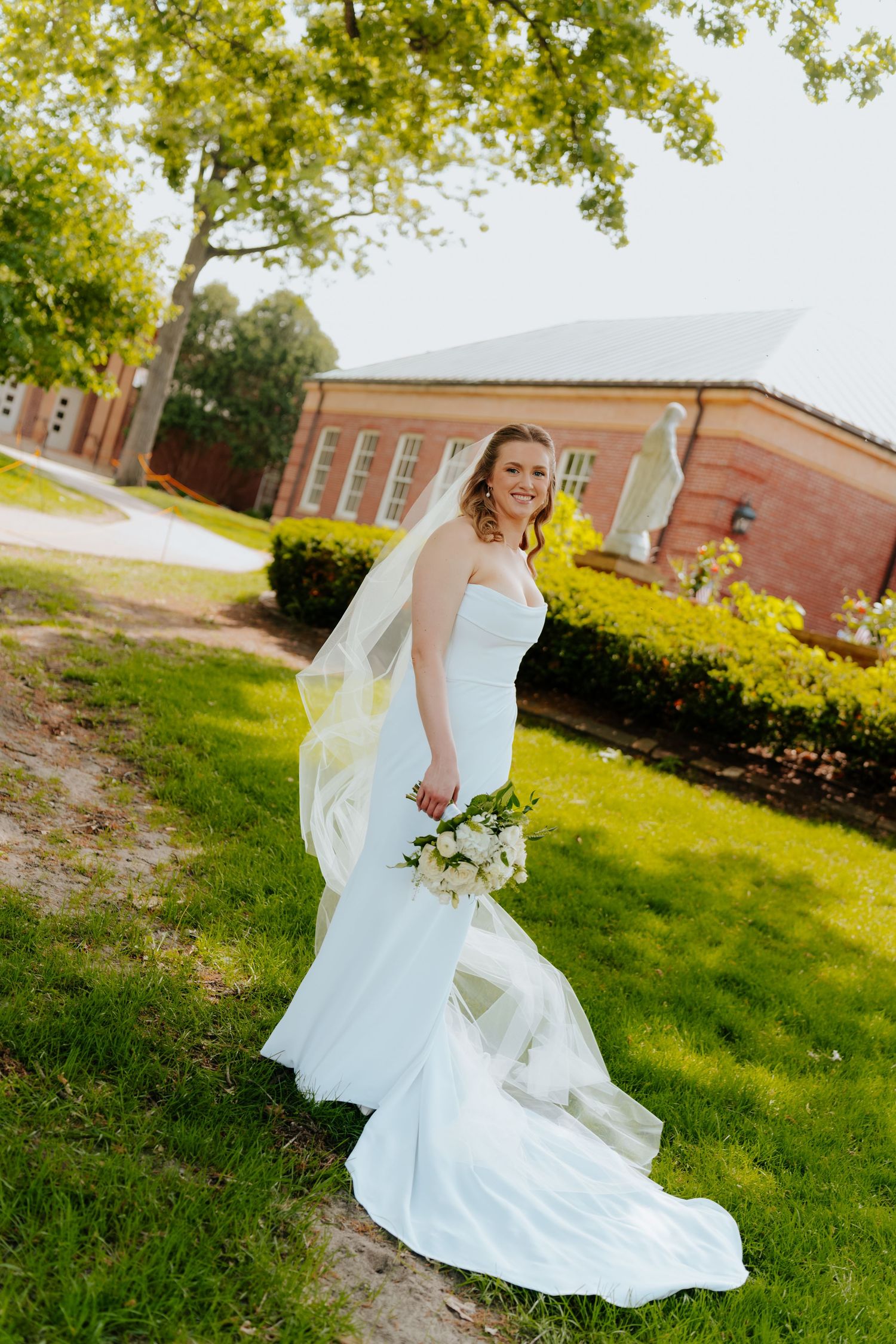 Smiling bride in white mermaid gown and veil holds bouquet outdoors near brick building on green lawn.