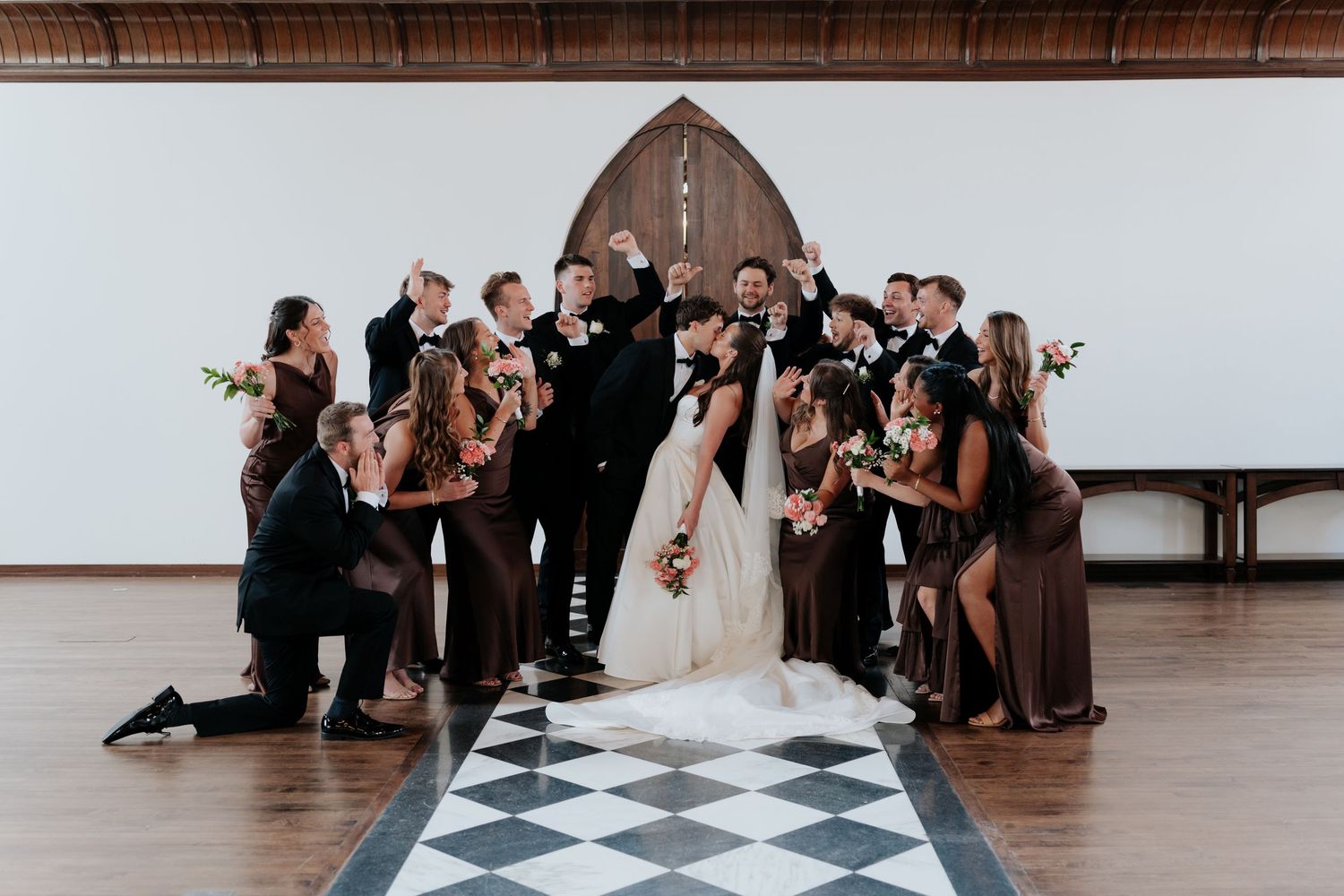 Wedding party celebrates bride and groom kissing on checkered dance floor in elegant venue.