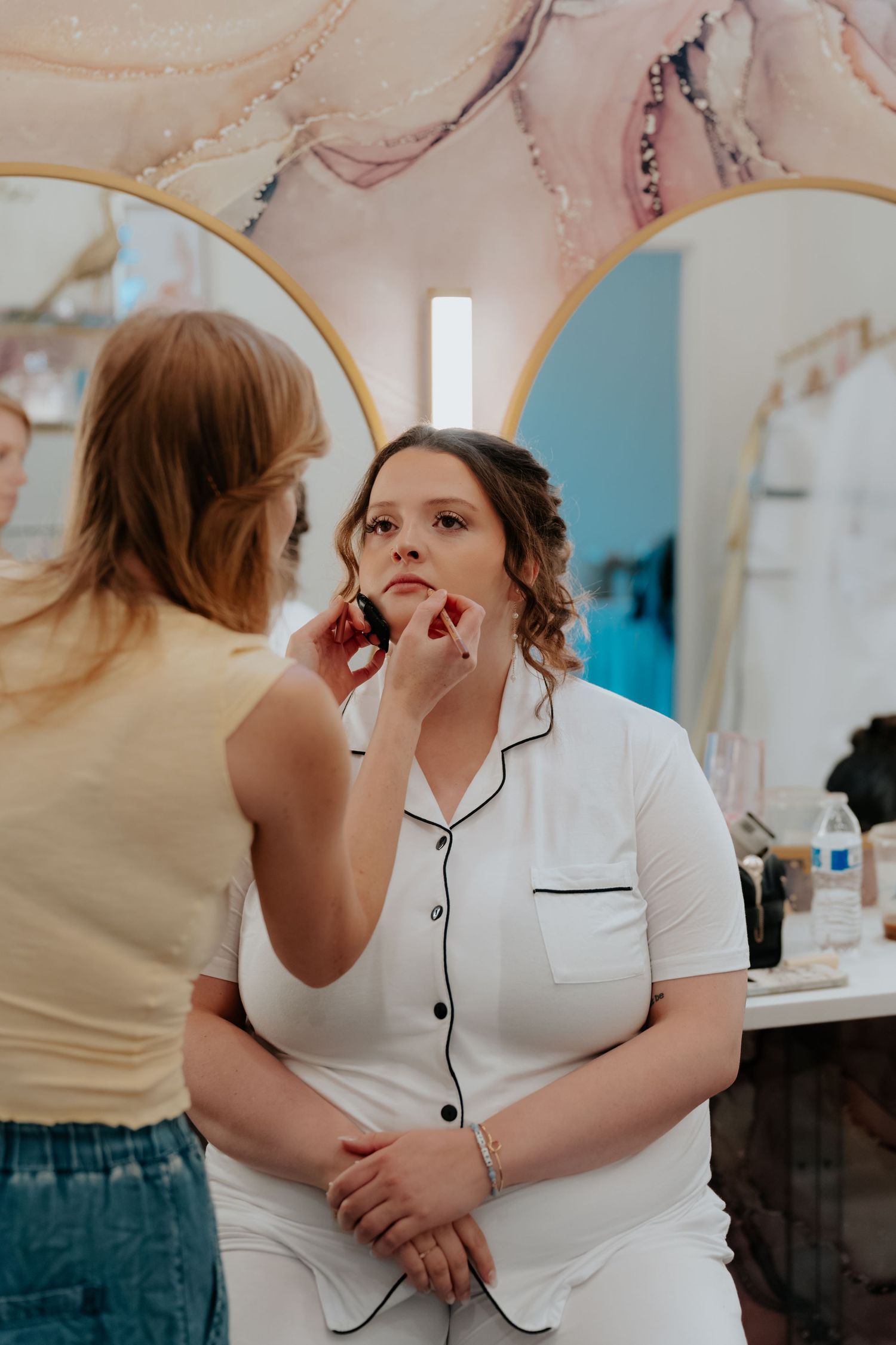 Makeup artist applying lipstick to a bride in white pajamas at a bridal suite vanity mirror.
