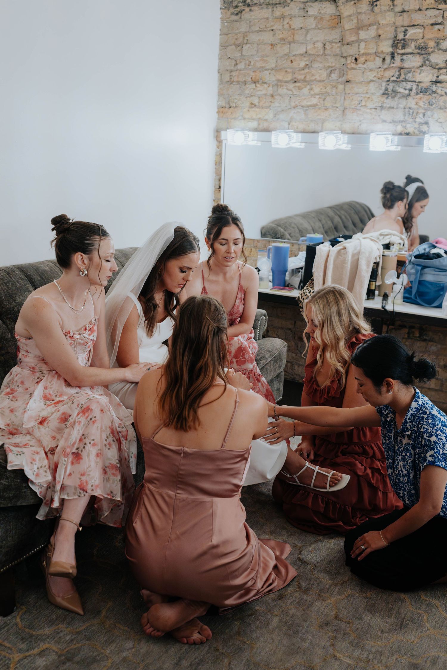 Bride in white veil sits with bridesmaids in floral and satin dresses, helping with wedding preparations in a rustic room.