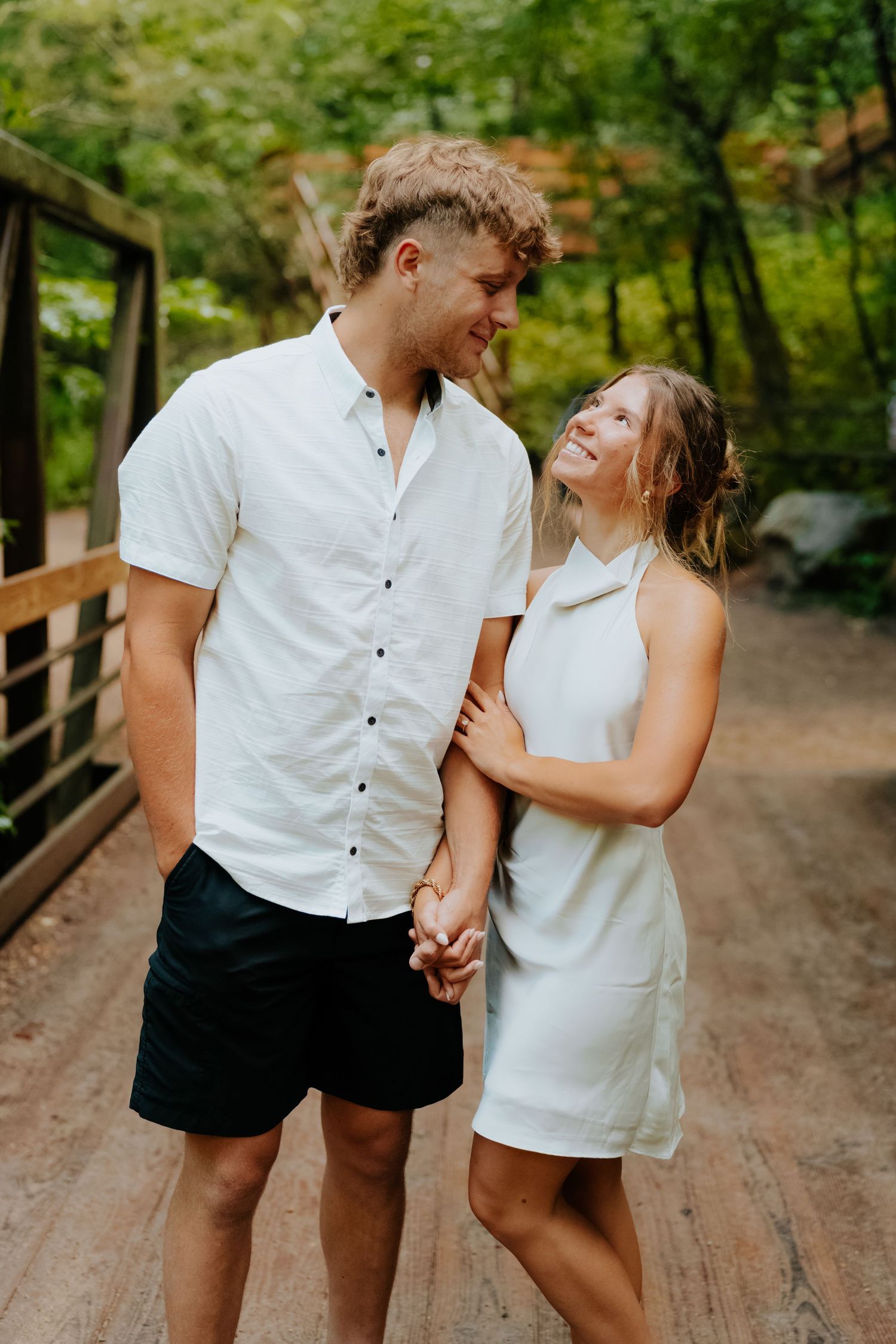 A couple in matching white outfits holds hands and smiles at each other on a wooden bridge surrounded by lush greenery.