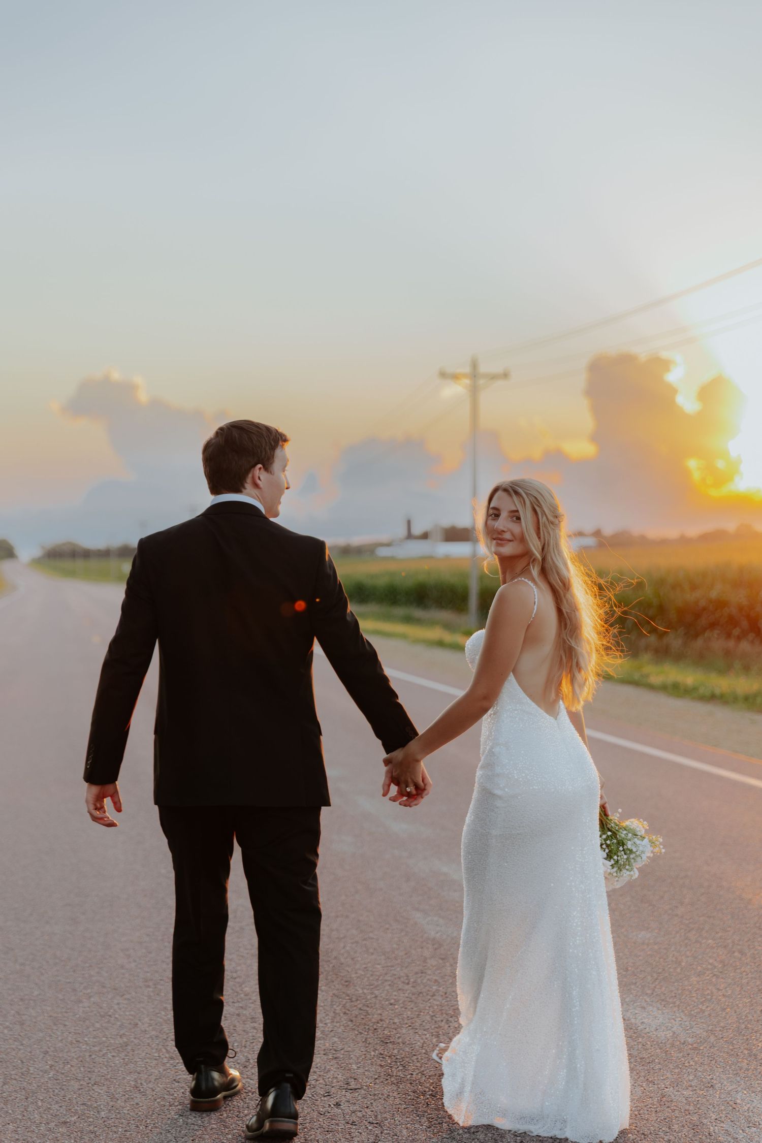 Newlyweds walking hand in hand on a country road during golden hour sunset.