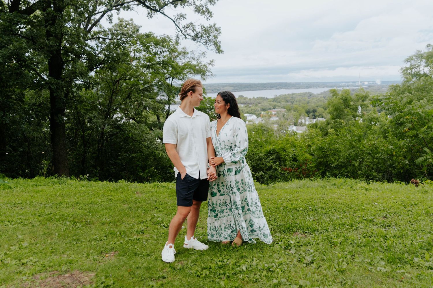 A couple shares a romantic moment on a hilltop overlooking a scenic landscape on a cloudy day.