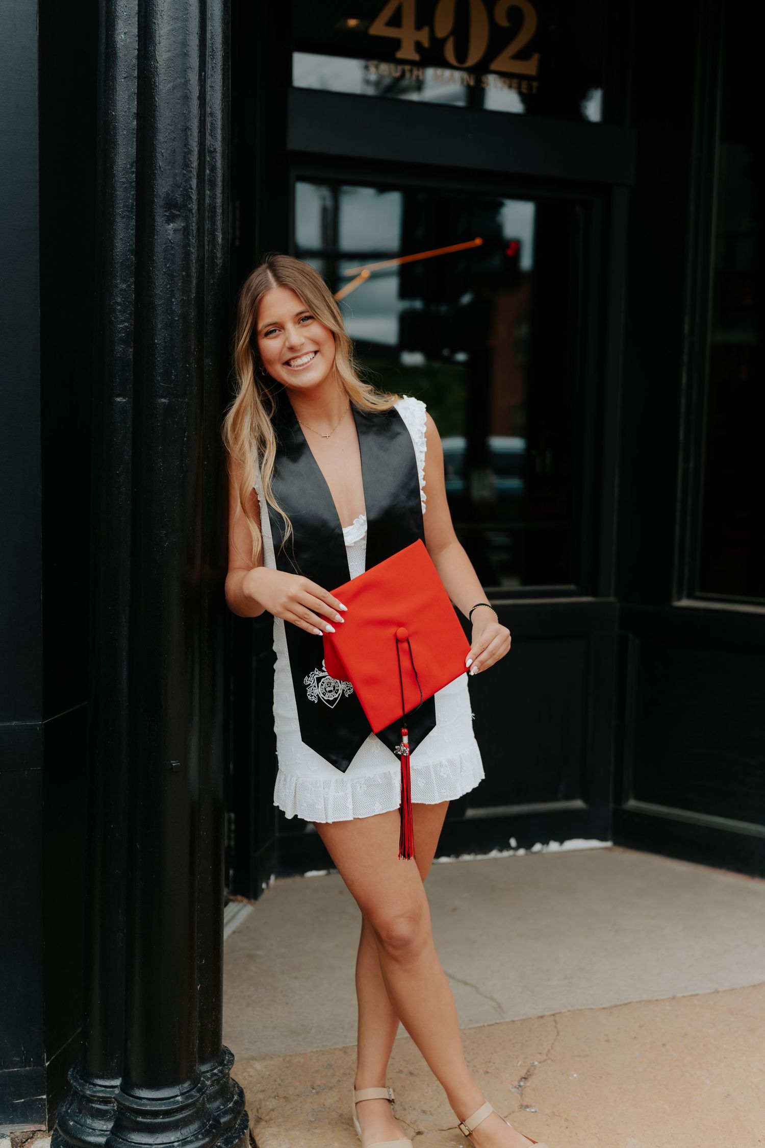 Smiling graduate in white dress holding red cap and wearing black stole, posing by pillar outside building 402.
