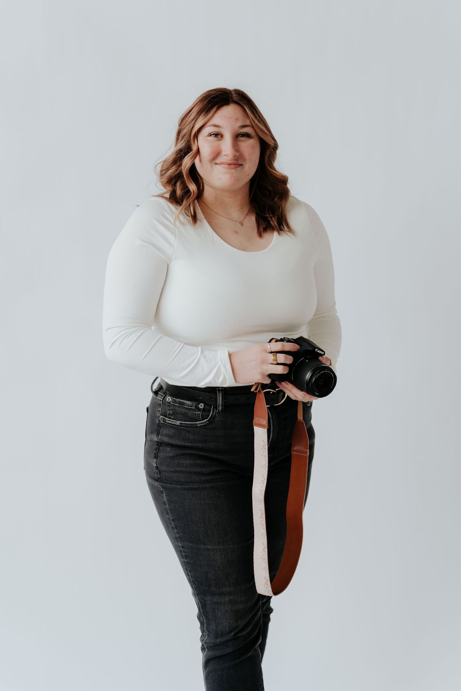 Smiling woman in white long-sleeve top and dark jeans holding a DSLR camera with brown strap on white background.