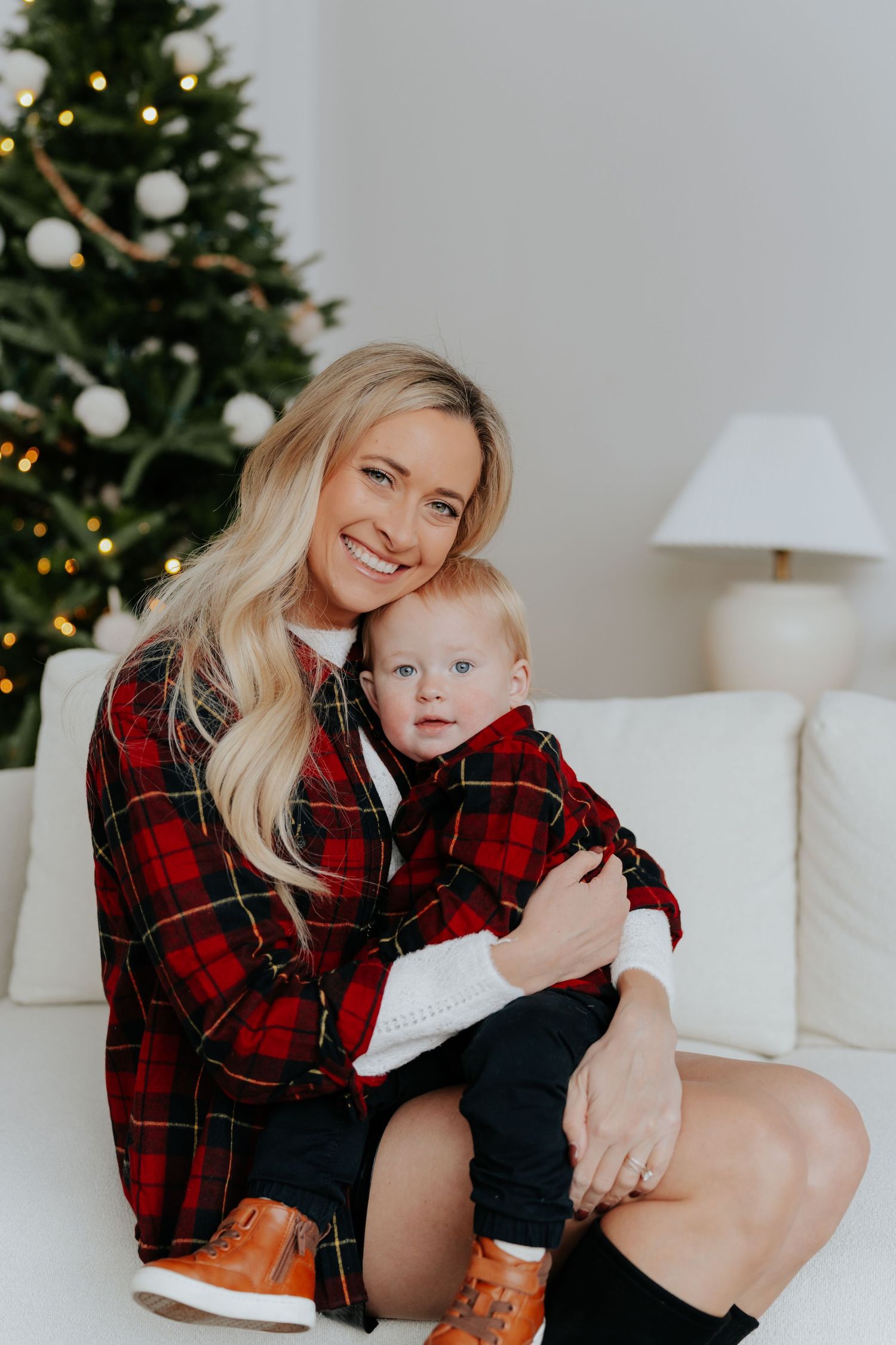 Smiling blonde woman in red plaid shirt holding baby in matching outfit near decorated Christmas tree.