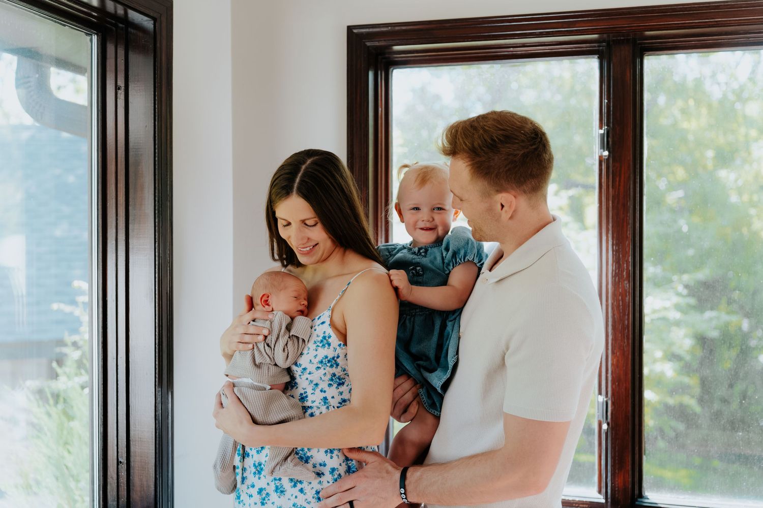 A young family with a newborn baby and toddler posing near bright windows in a modern home.