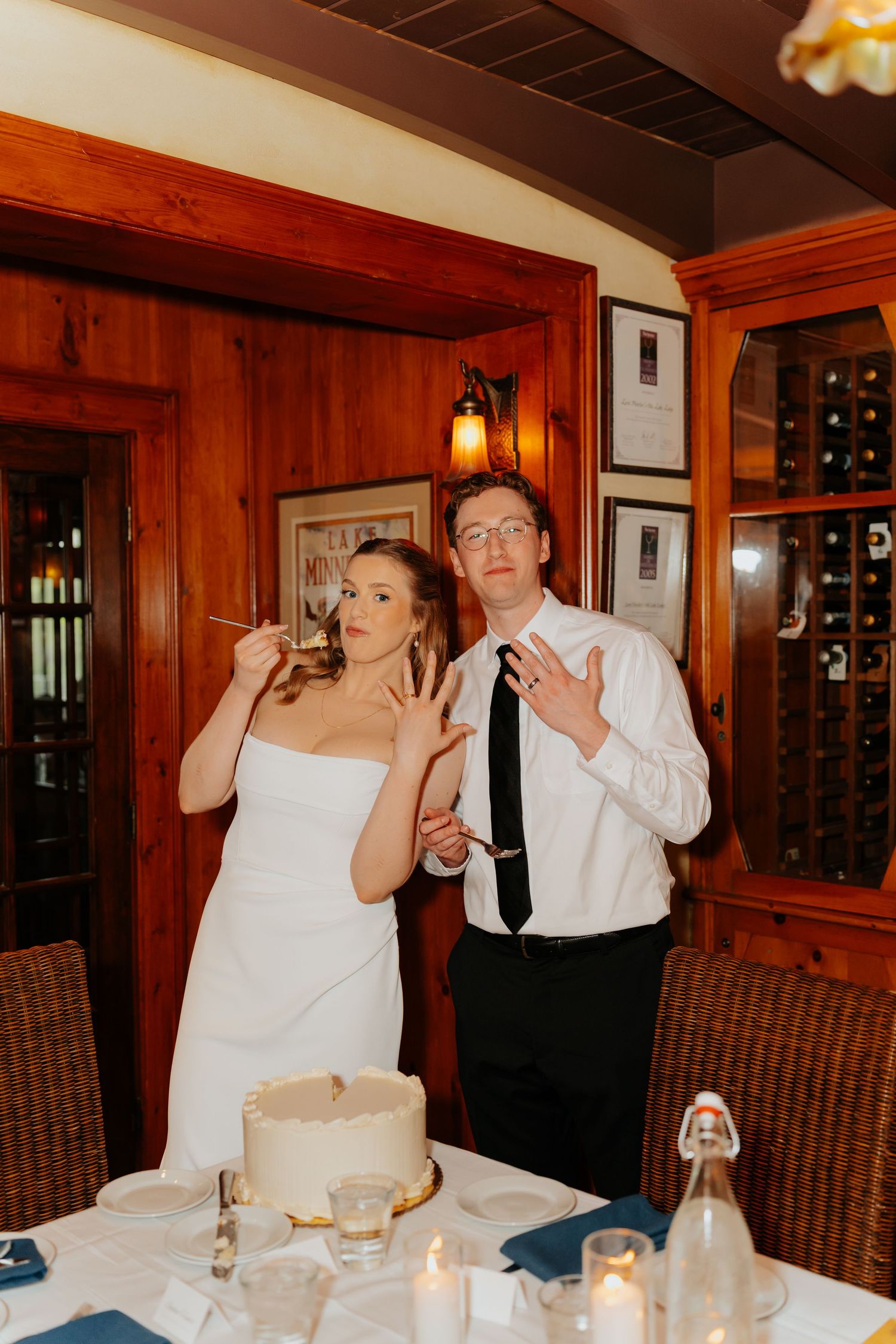 Newlyweds show off wedding rings beside cake, bride in white gown, groom in white shirt and black tie, in a wood-paneled restaurant.