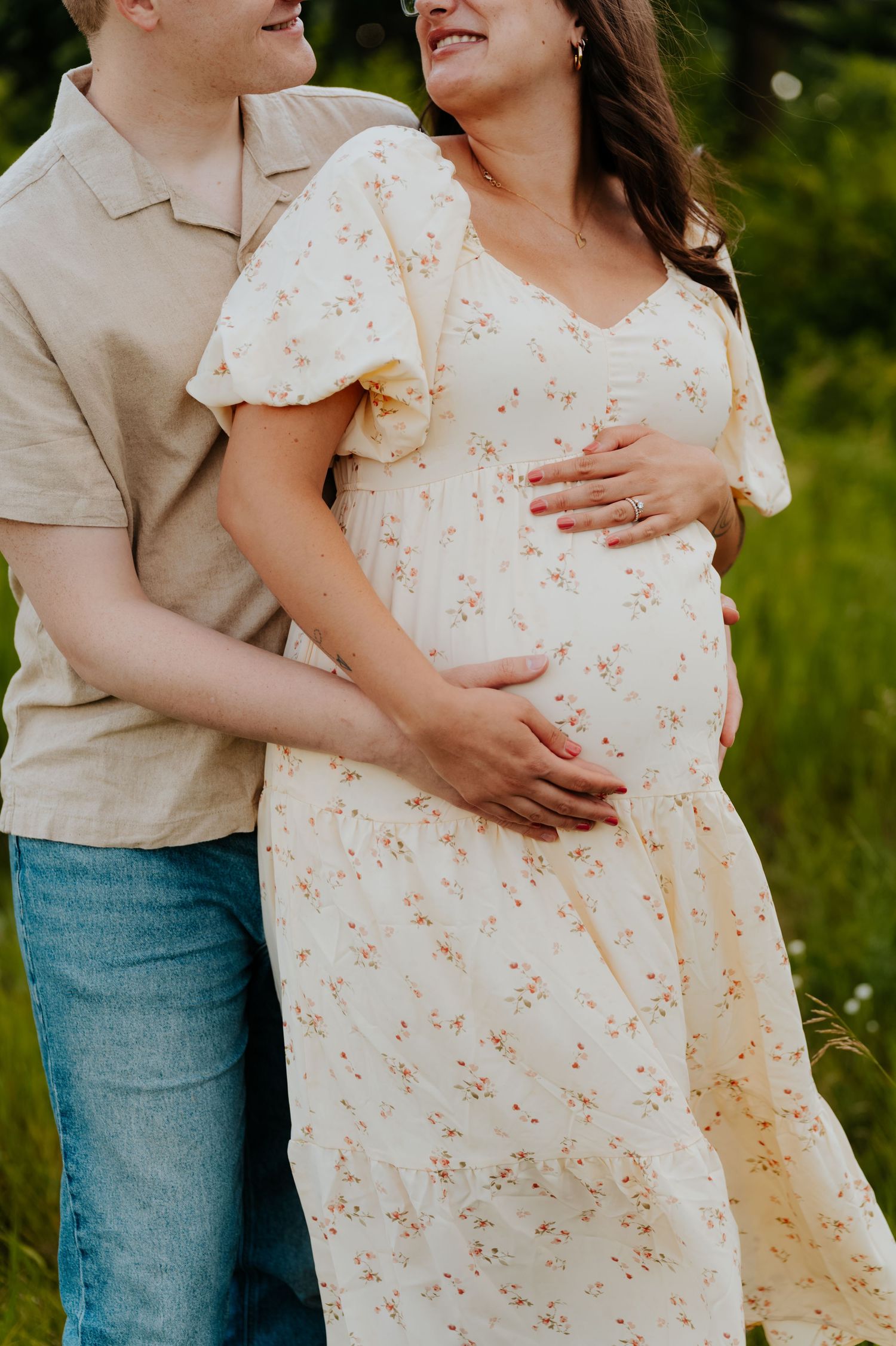 Couple embracing outdoors, pregnant woman in floral dress with partner holding her baby bump in a green field.