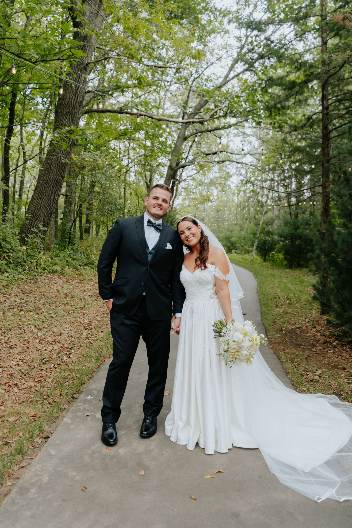 Bride and groom posing on a tree-lined path, bride in white gown holding bouquet, groom in black tuxedo.