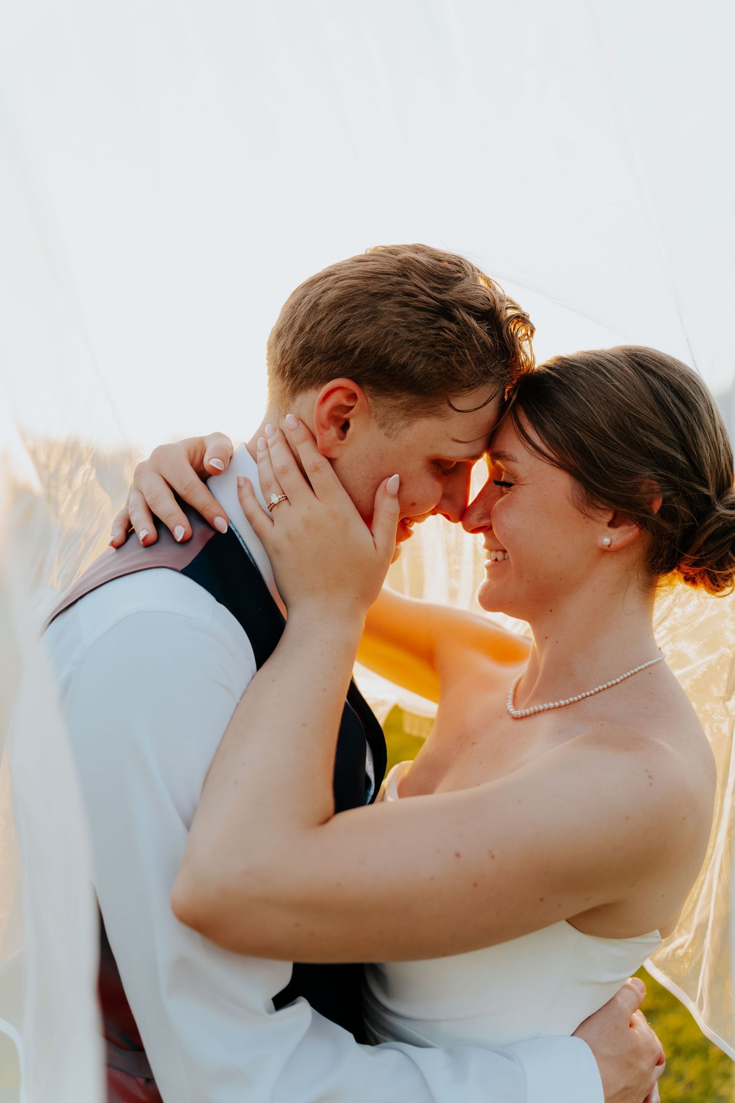 A bride and groom share a romantic forehead touch under a flowing veil, bathed in warm golden sunset light.