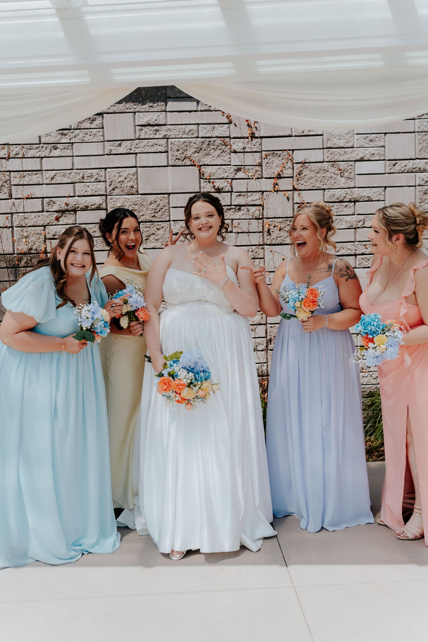 Bride in white gown laughing with four bridesmaids in pastel dresses holding colorful bouquets at wedding venue.