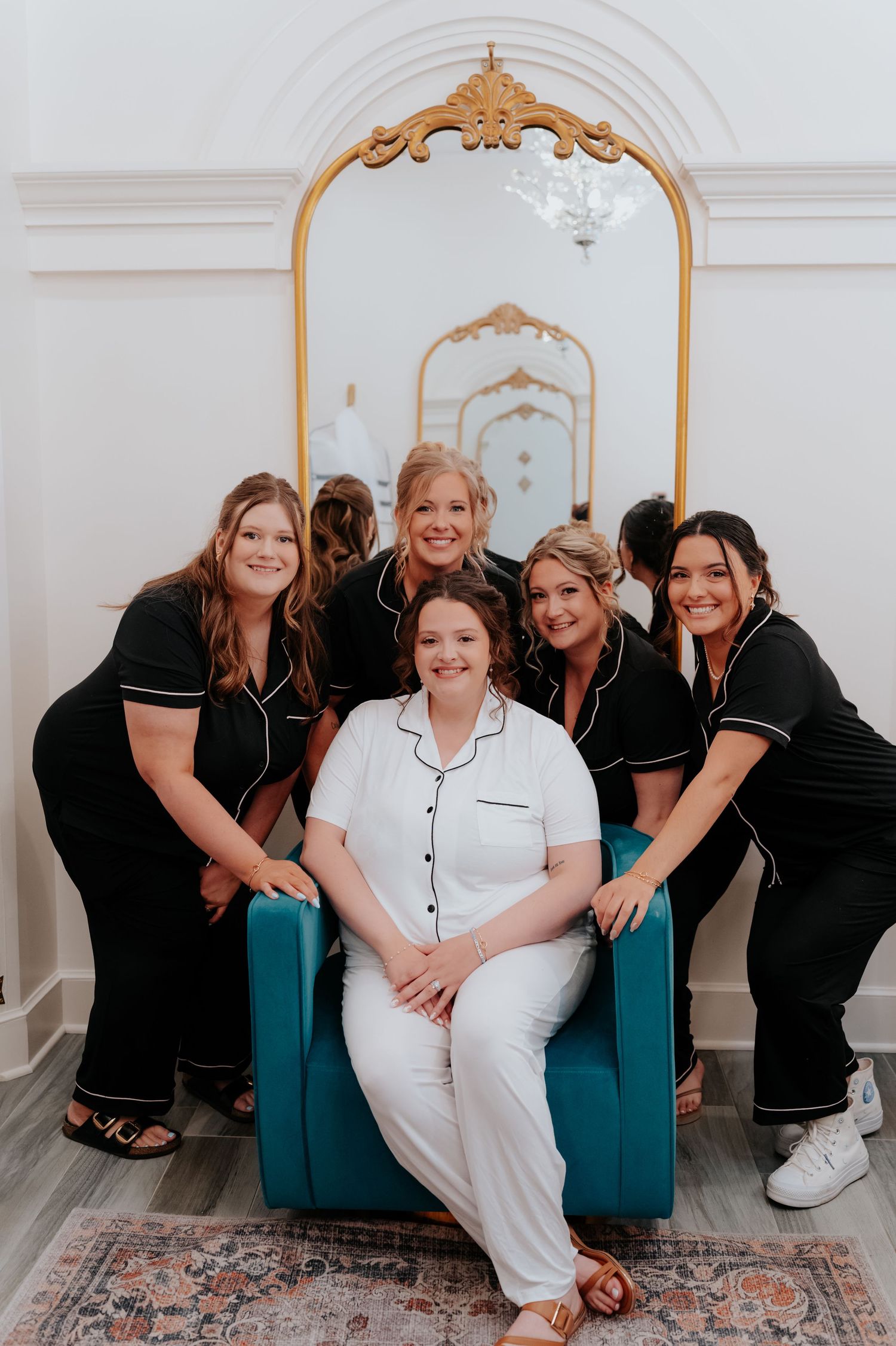 Bride in white pajamas sits in teal chair surrounded by bridesmaids in black pajamas before wedding day preparations.