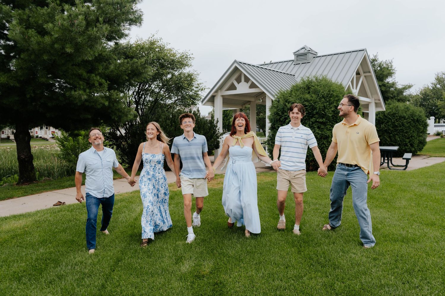 A joyful family of six holds hands walking on green grass near a white cottage surrounded by trees.