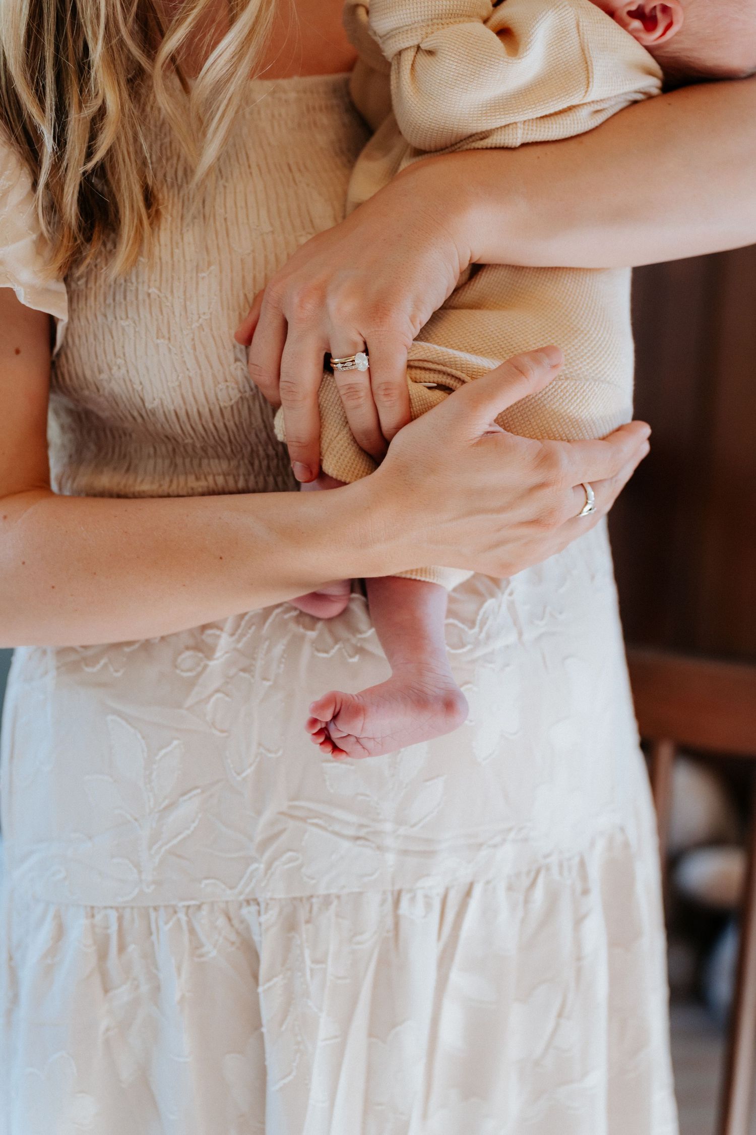Mother in white dress tenderly holding newborn baby, showcasing tiny feet and wedding rings in soft natural light.
