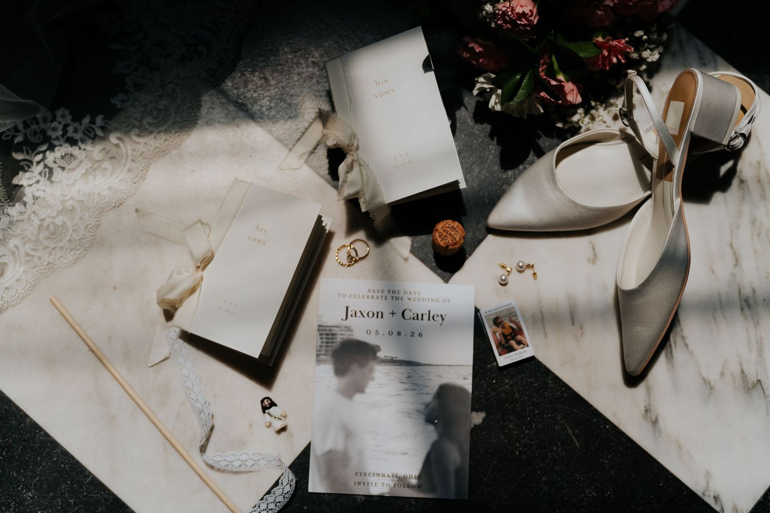 Elegant wedding flat lay with invitation, white heels, rings, and floral bouquet on marble surface.