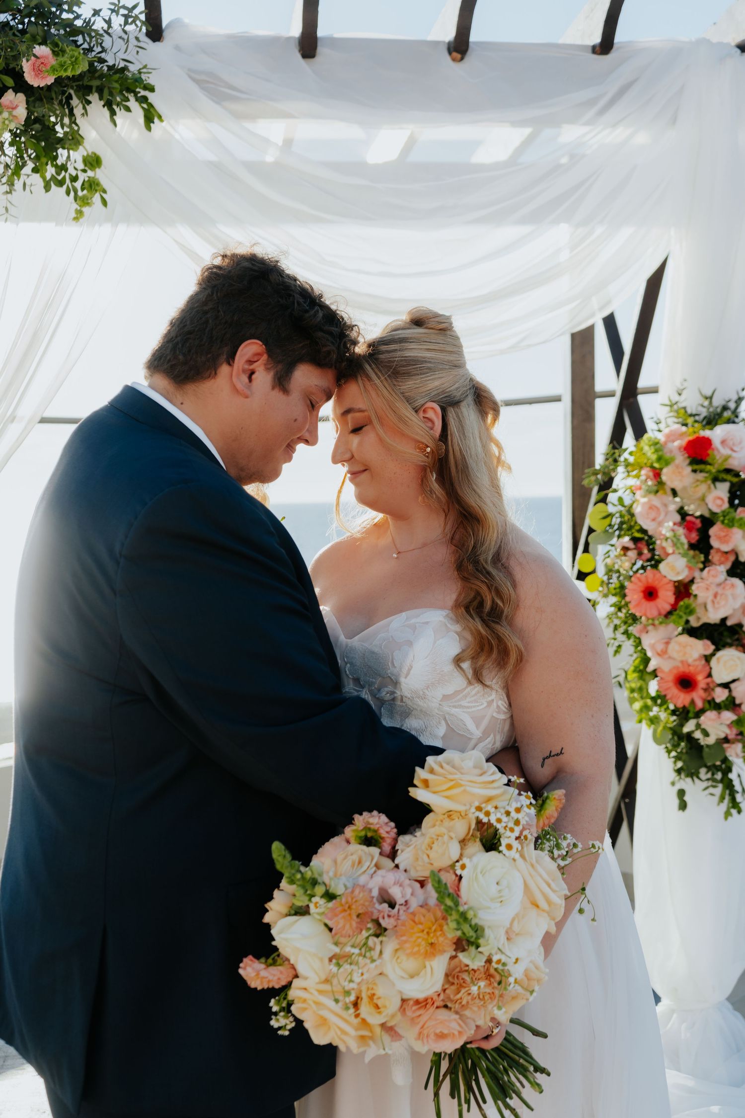 Bride and groom touching foreheads under floral arch at outdoor wedding ceremony with white draping.
