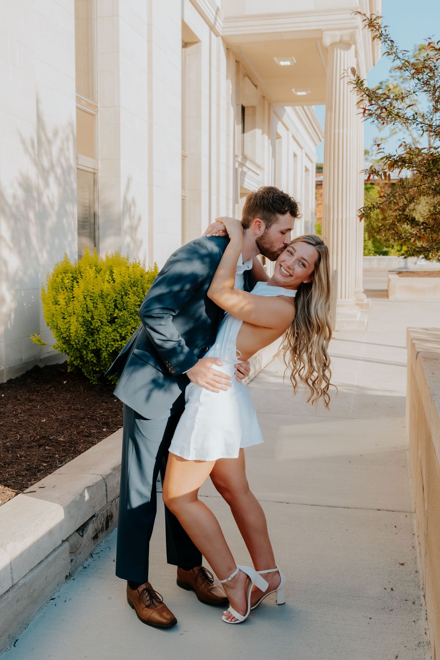 Couple embracing and laughing outside elegant white building with columns during engagement photo session.