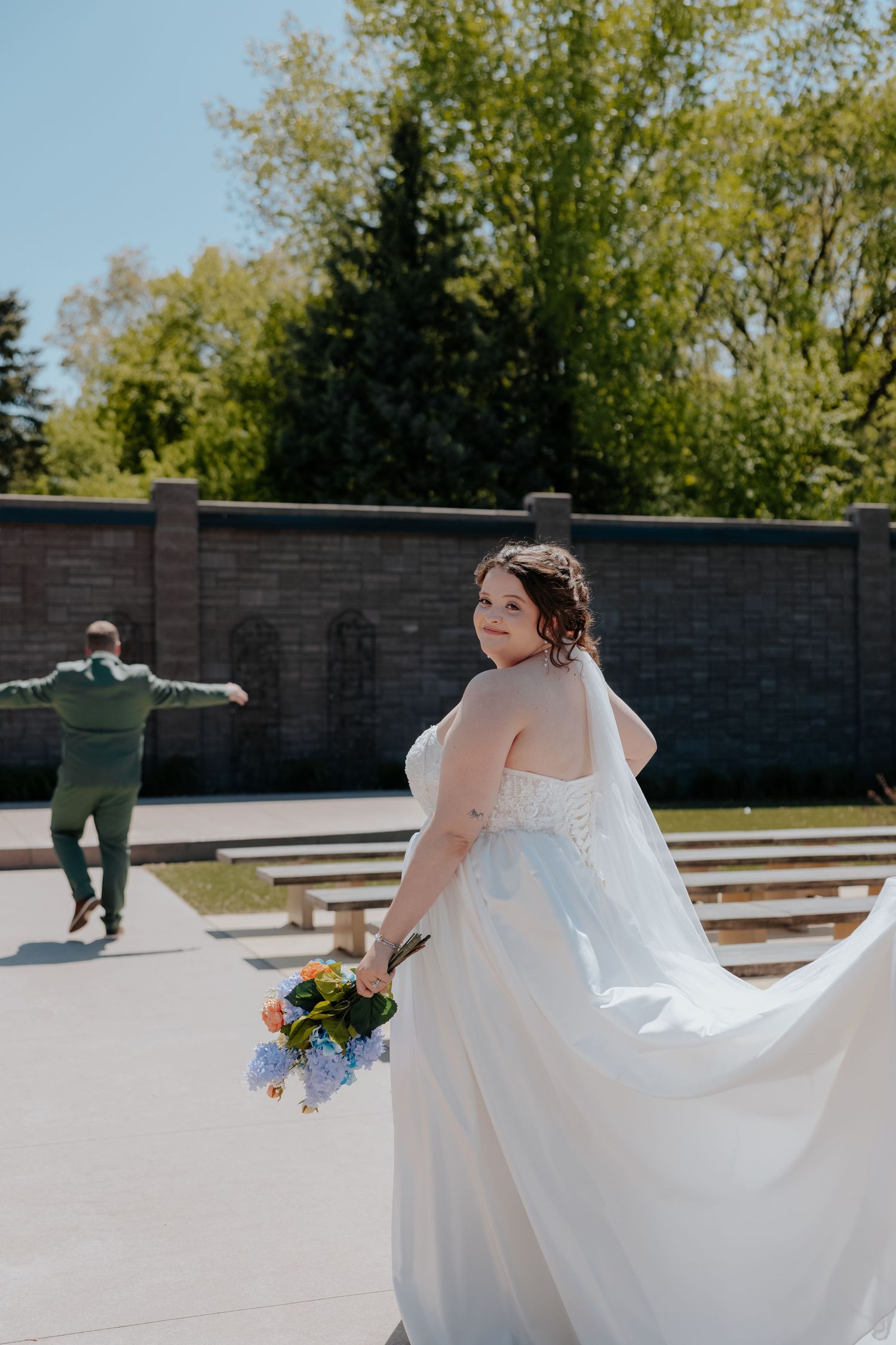 Smiling bride in white gown holding bouquet as groom runs toward her outdoors.