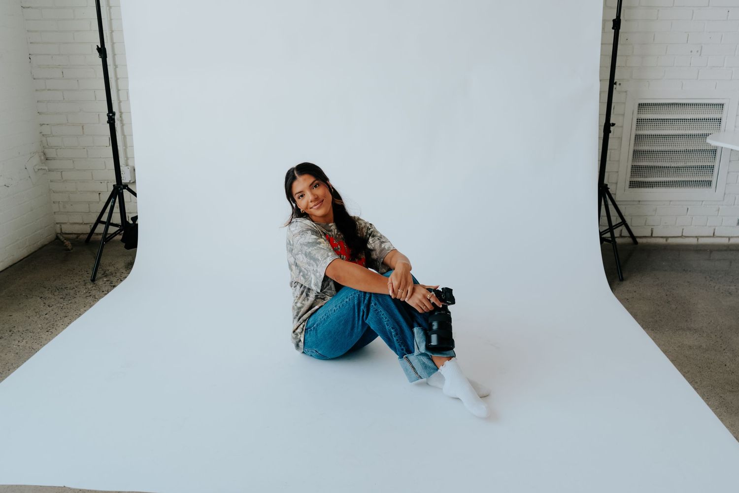 Woman sitting on floor against white backdrop in photography studio, wearing jeans and patterned shirt, holding camera.