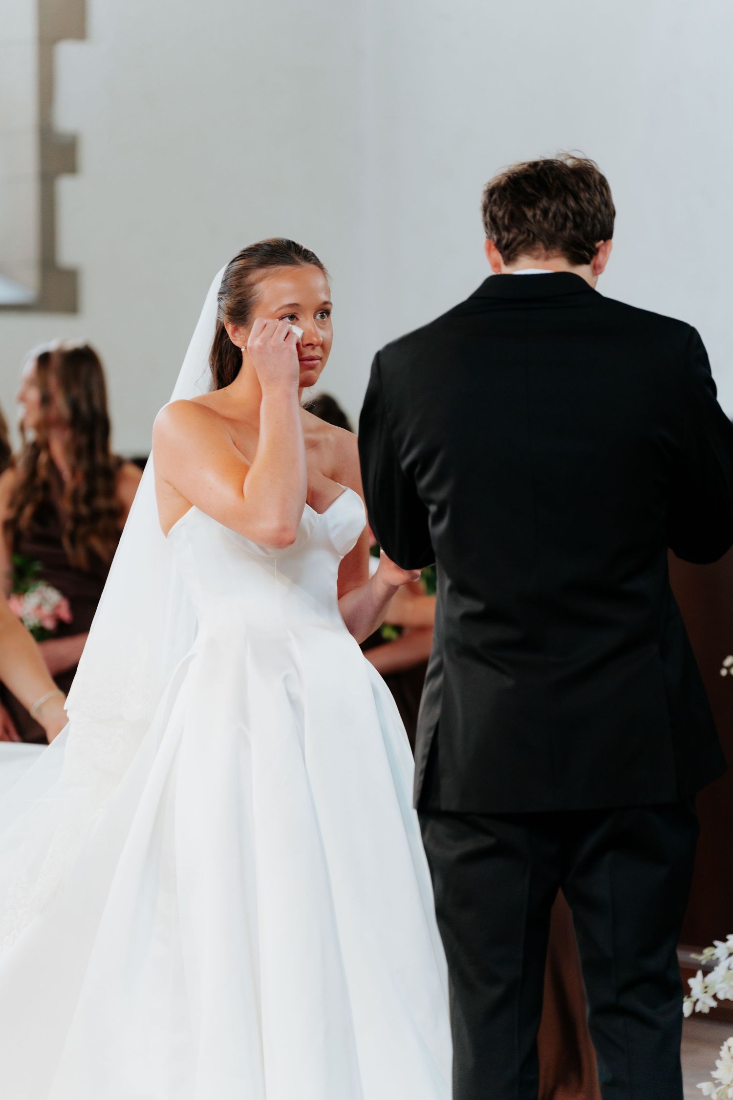 Emotional bride in white gown wiping tears during wedding ceremony while groom stands before her.