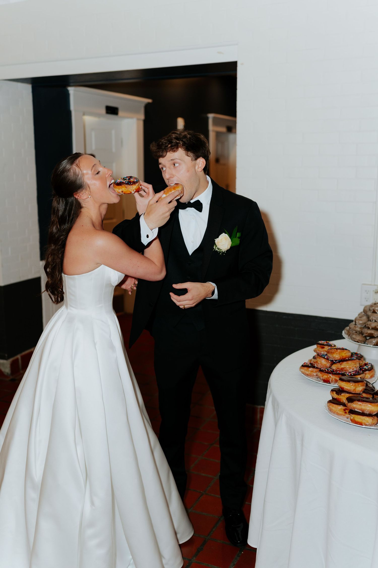Bride feeds groom a chocolate donut at their wedding reception, with a display of pastries nearby.