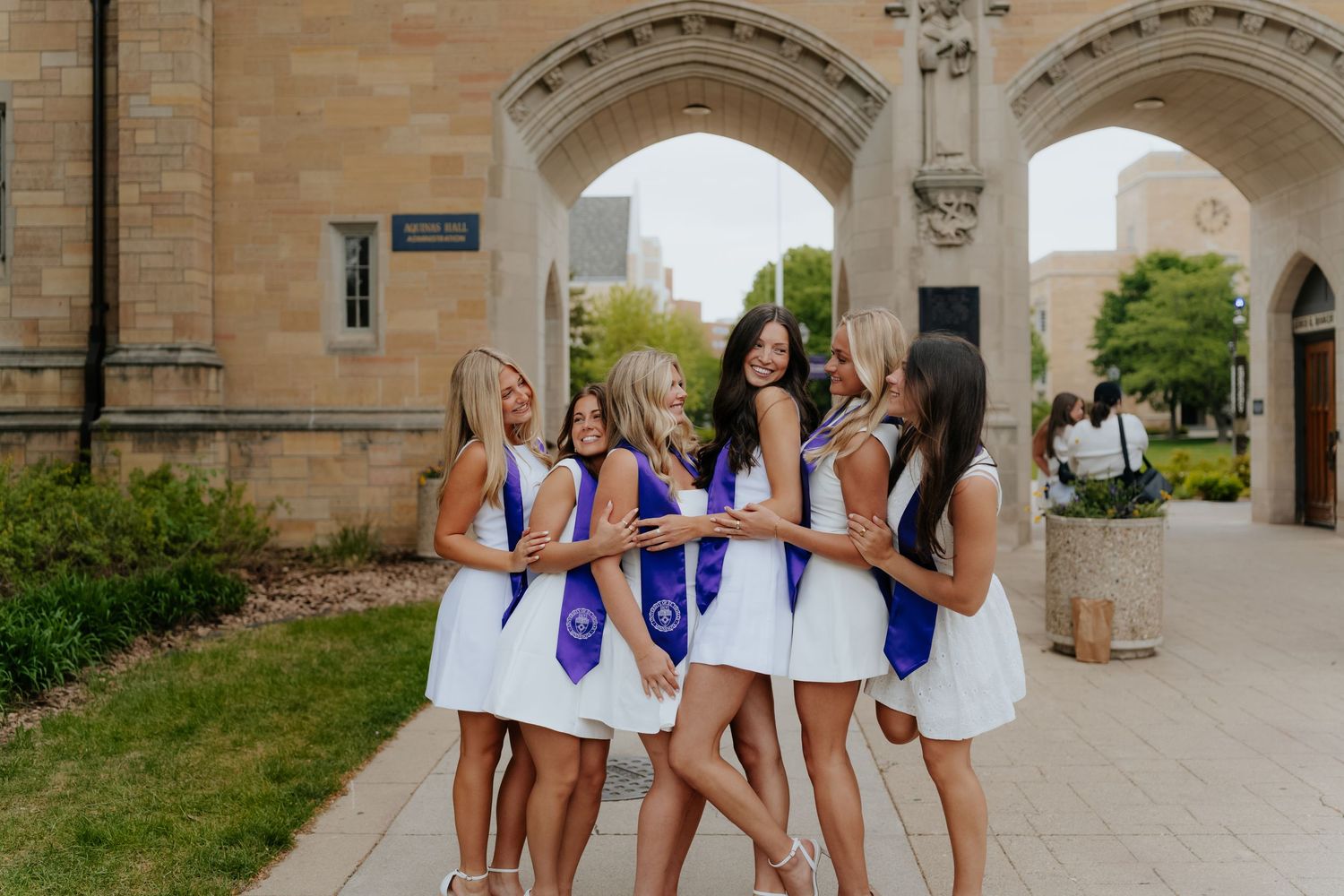 Six female graduates in white dresses and purple sashes celebrate together outside a stone university building.