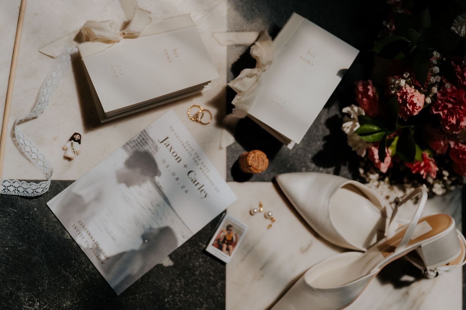 Elegant wedding flat lay with white stationery, bridal heels, gold rings, floral bouquet, and ribbon on marble surface.