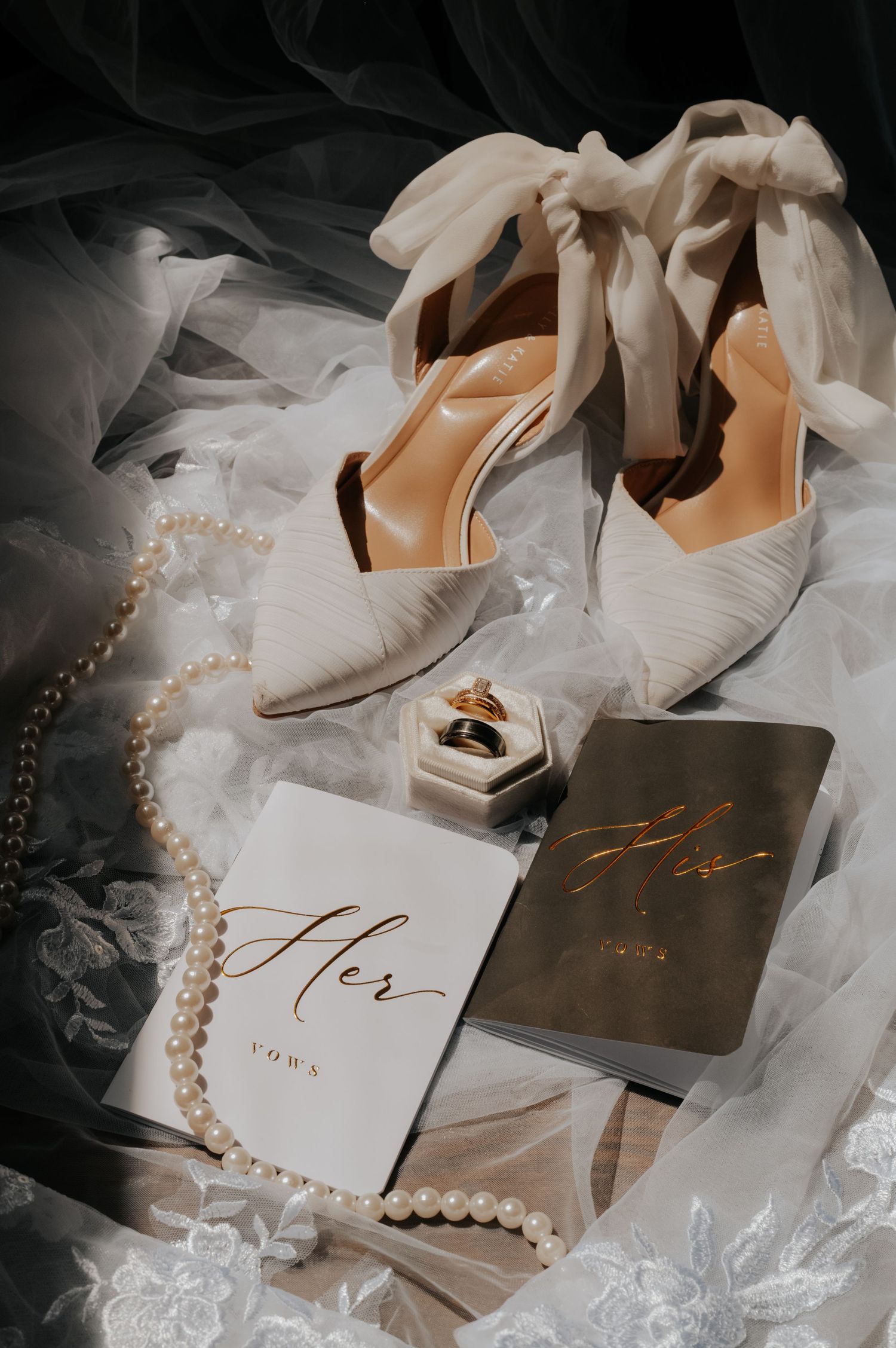Elegant bridal flat lay with white heels, pearl necklace, gold ring, and wedding vow books on lace fabric.
