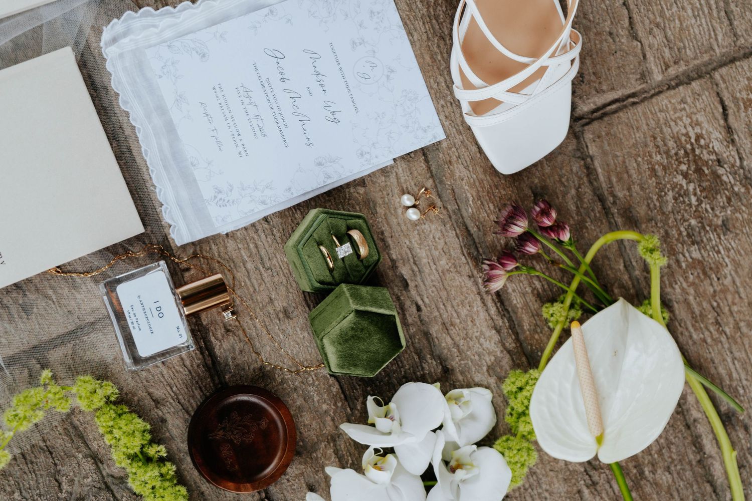 Elegant wedding flat lay with invitation, white heels, green hexagon ring box, perfume, and white anthurium flowers on wood.