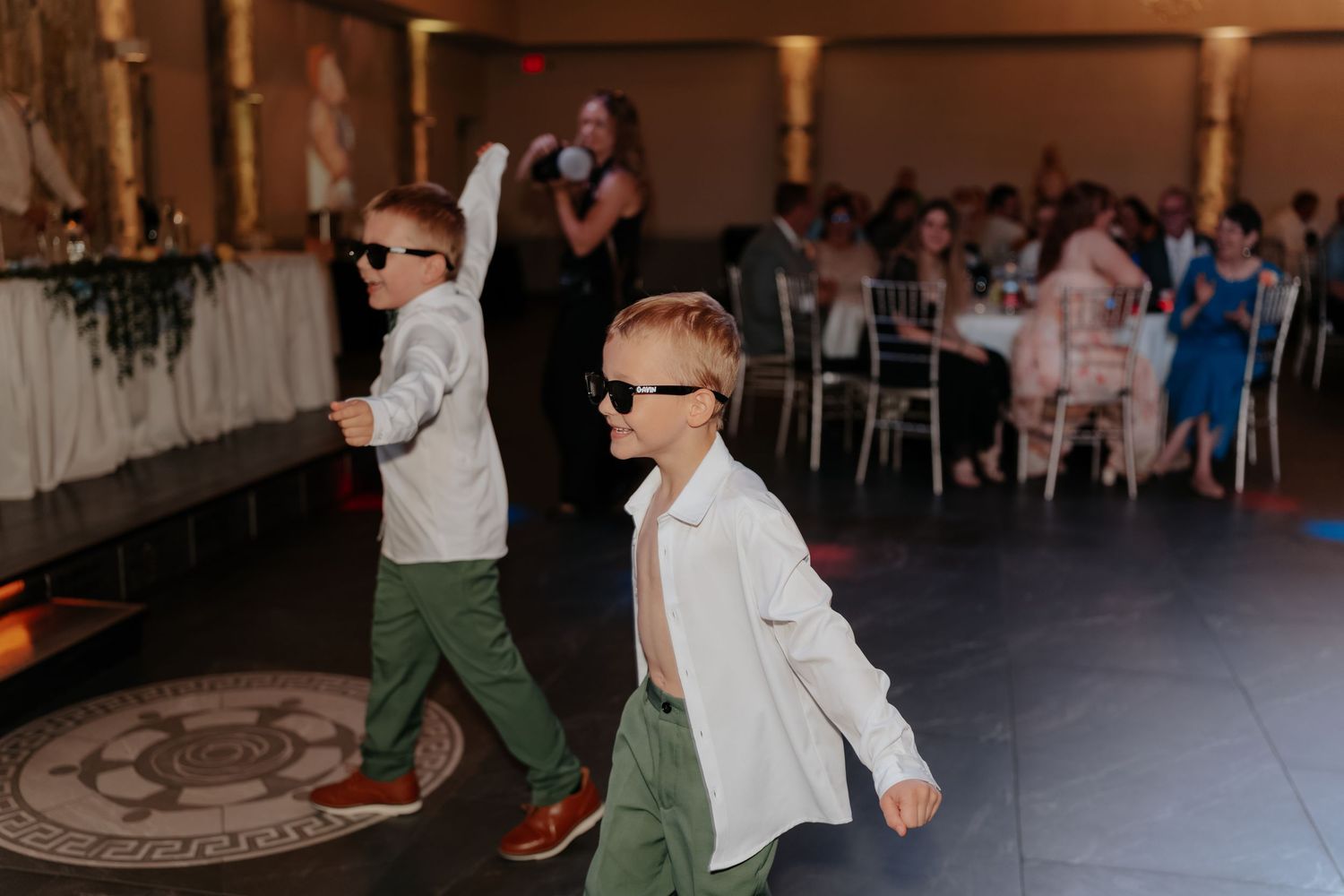 Two young boys in sunglasses and green pants dance at a wedding reception on a decorated dance floor.