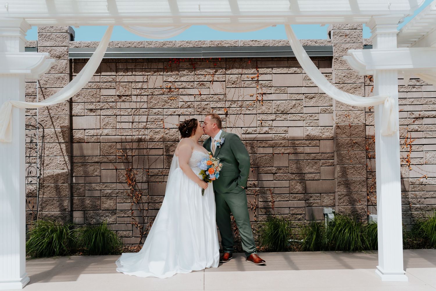 Bride and groom sharing a kiss under a white pergola with draped fabric at an outdoor wedding ceremony.