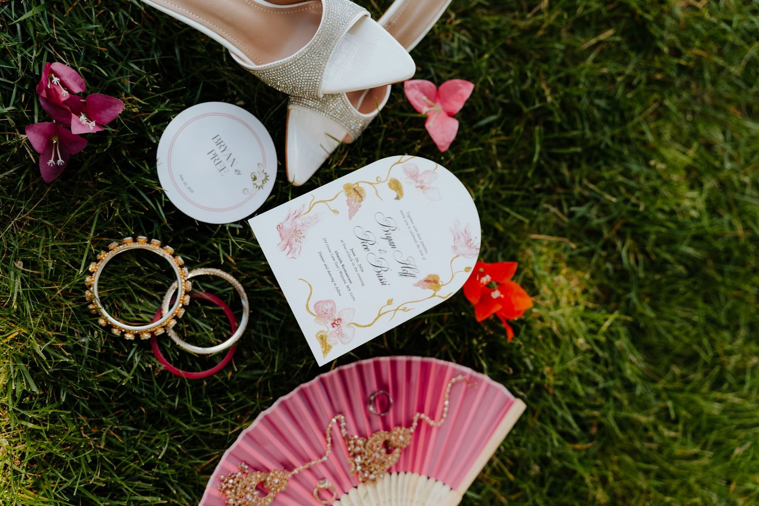 Wedding flat lay on grass featuring bridal shoes, pink fan, gold rings, floral invitation, and colorful flowers.