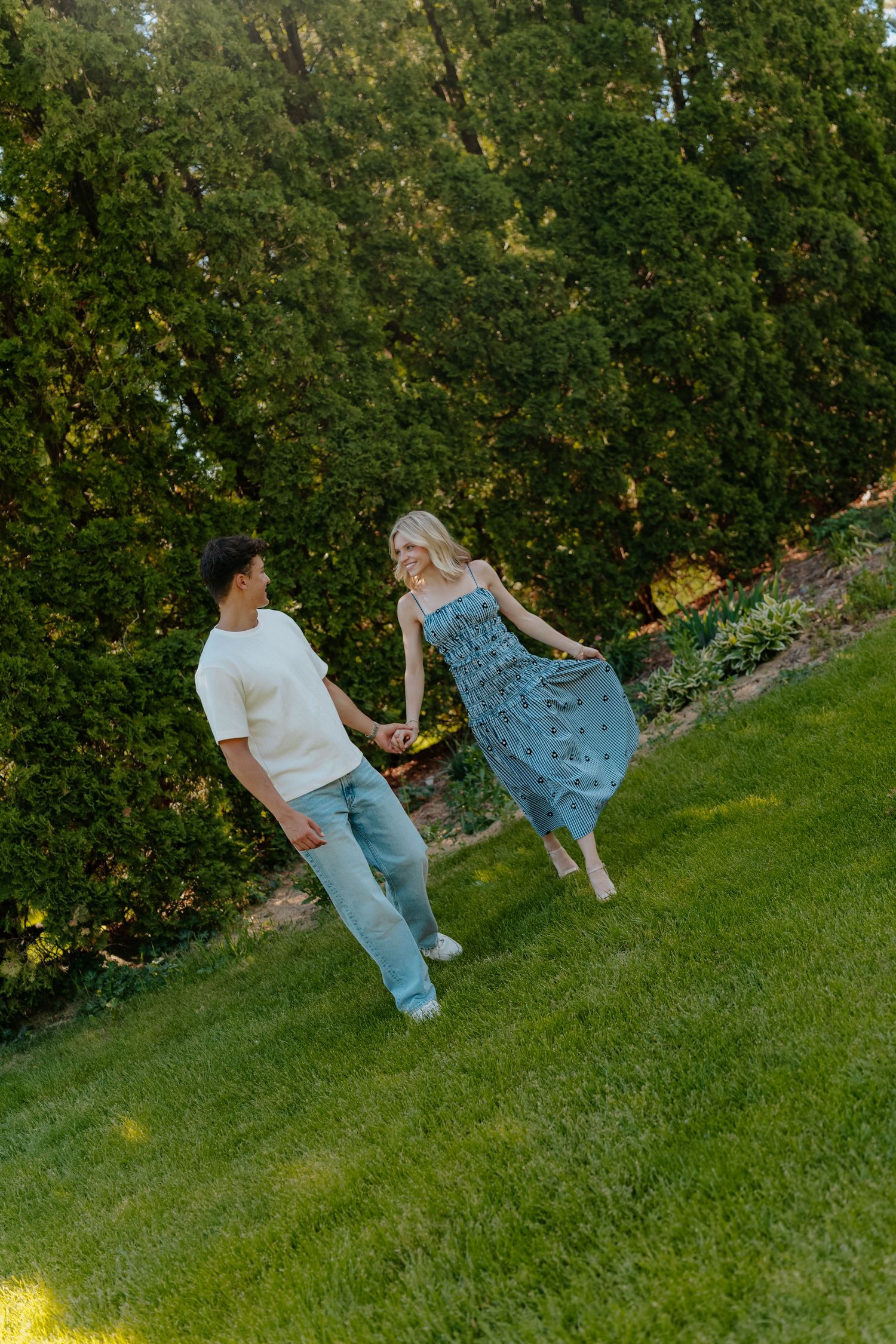 A couple holds hands while walking on lush green grass surrounded by tall evergreen trees in a garden.