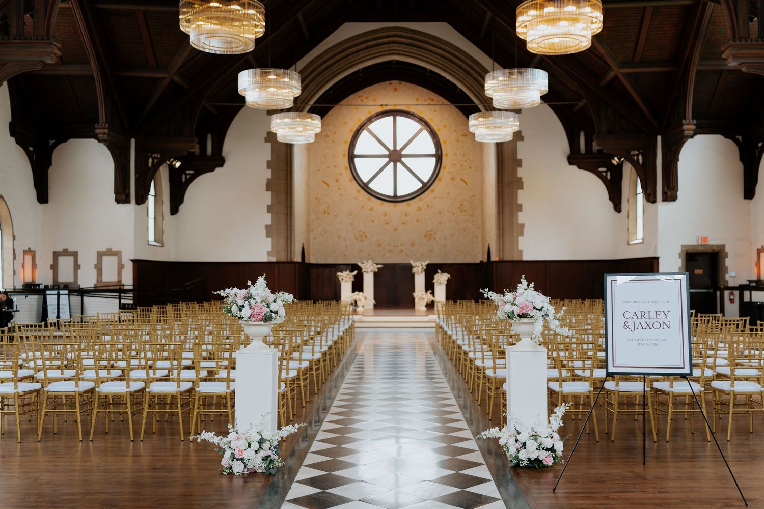 Elegant wedding ceremony setup with gold chairs, floral arrangements, and a checkered floor in a grand hall.