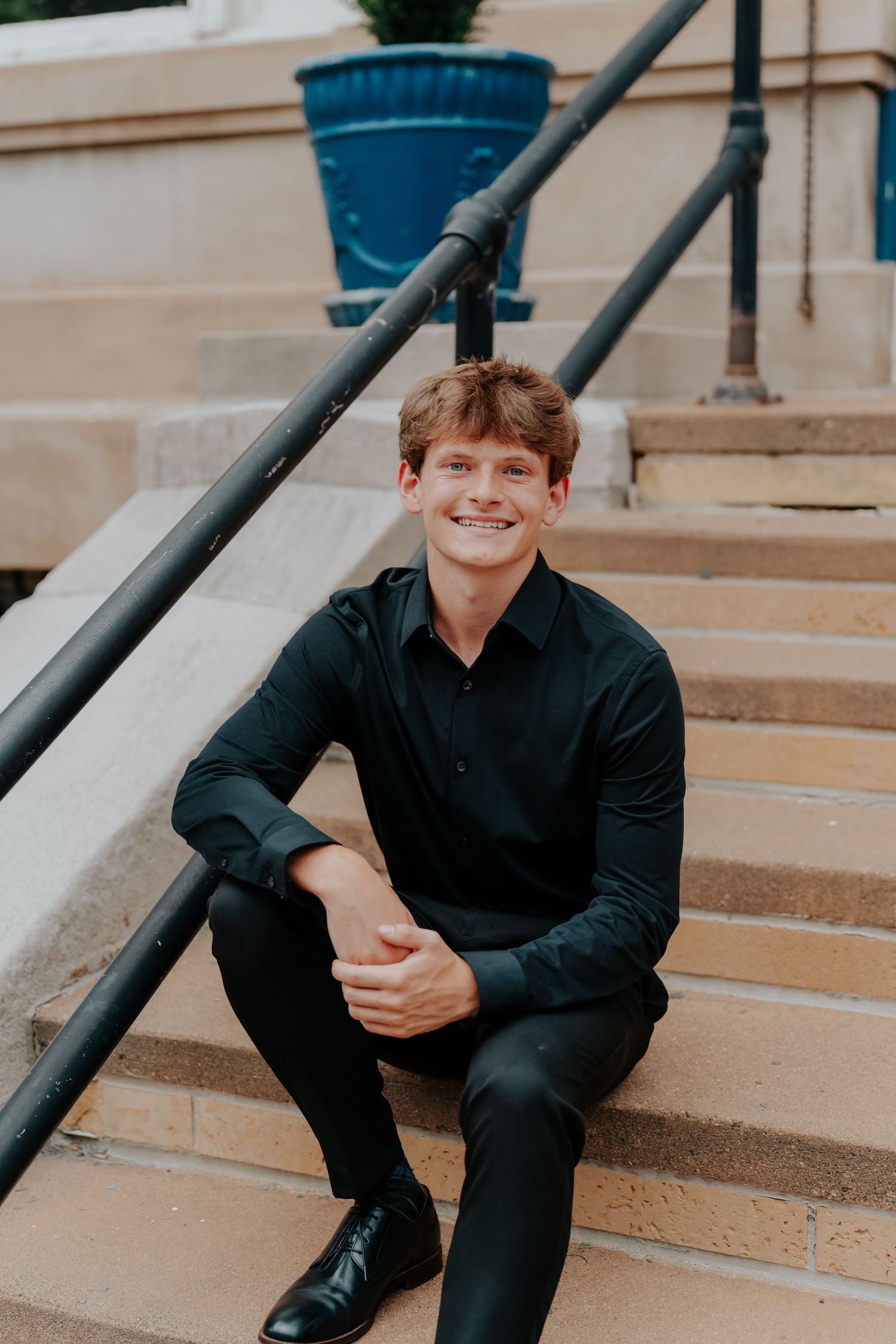 Young man in black outfit smiling on outdoor stone steps, with metal railing and blue planter in background.