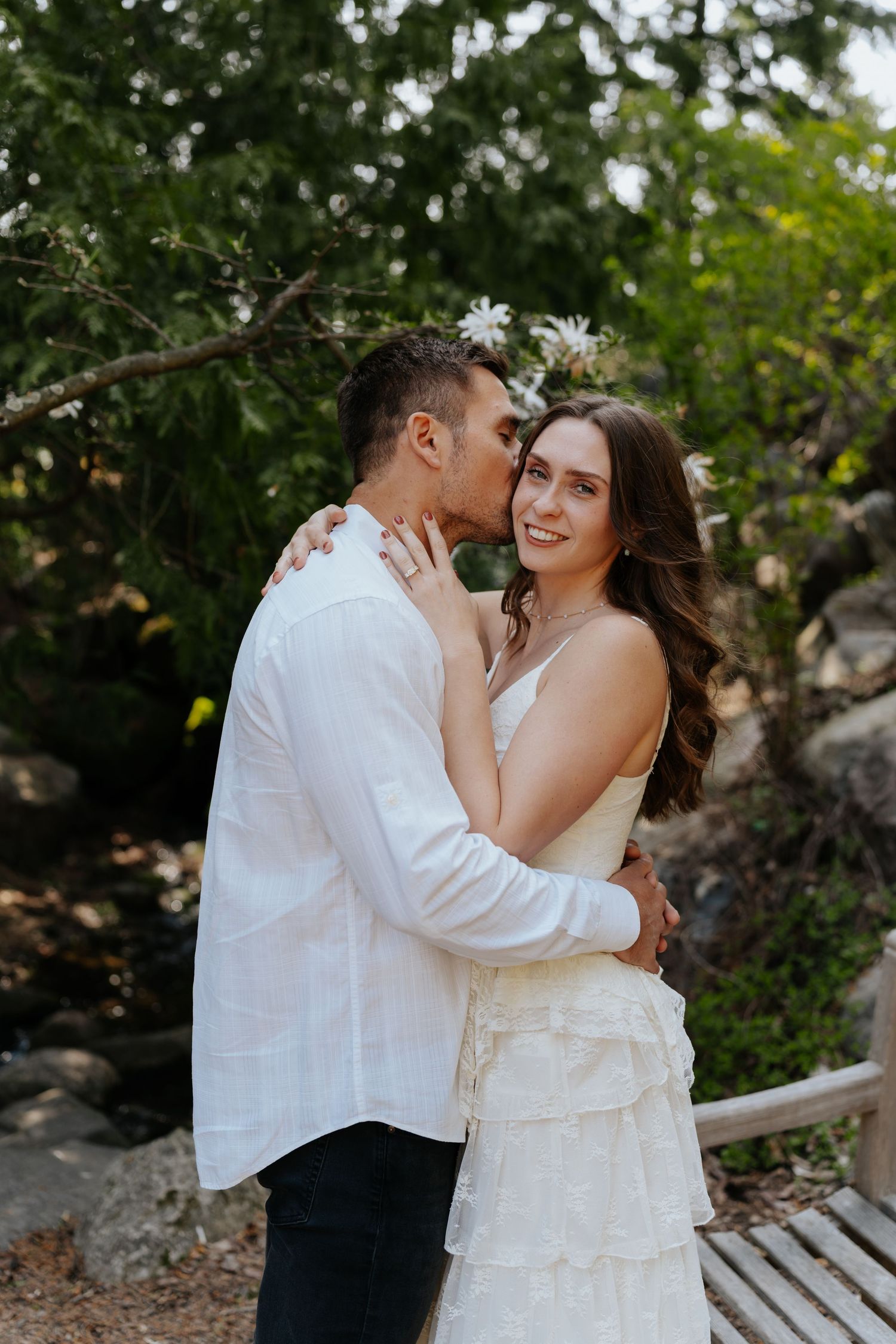Couple embracing in garden, man kissing woman's cheek as she smiles, both wearing white near lush greenery.