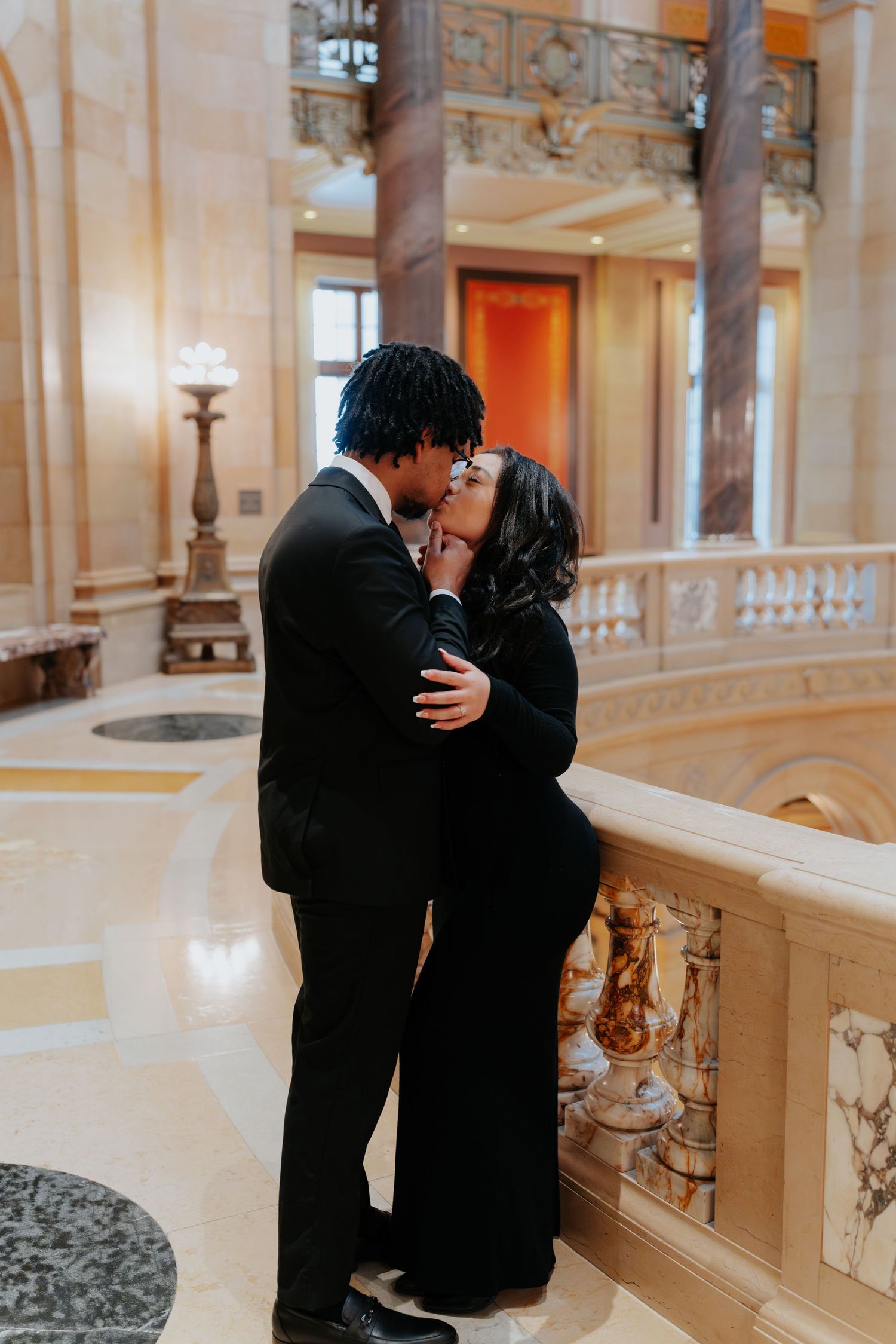 Couple dressed in black sharing a romantic kiss in a grand marble building interior.
