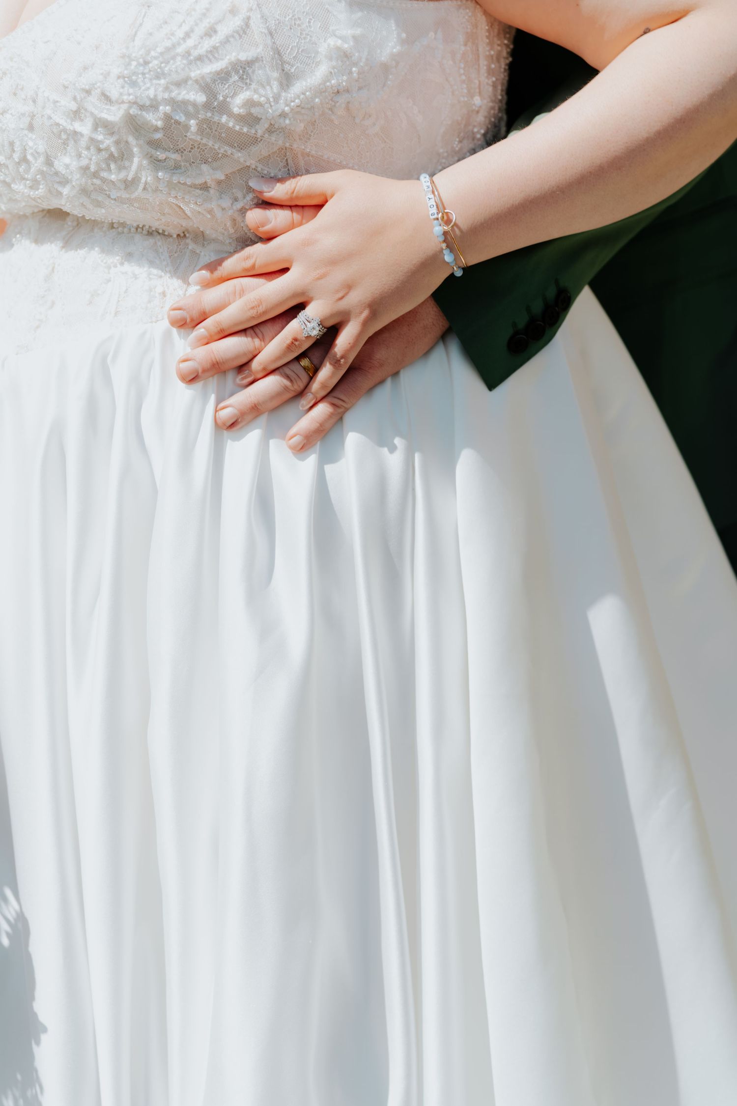 Close-up of bride and groom hands with diamond ring and bracelet on white wedding gown.