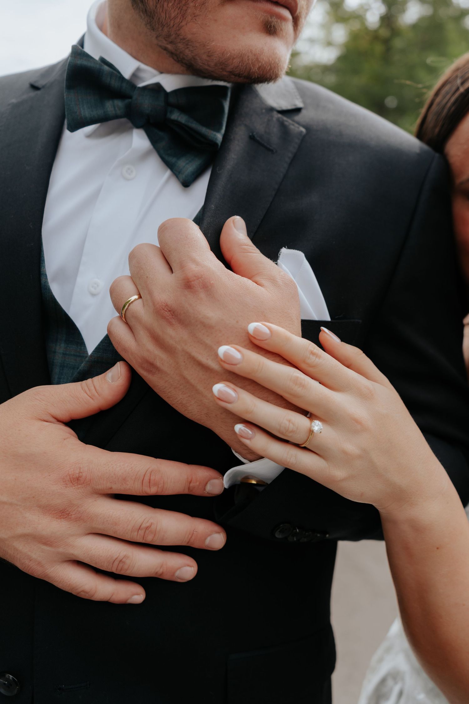 Bride and groom close-up showing wedding rings, white manicure, black tuxedo with green bow tie.