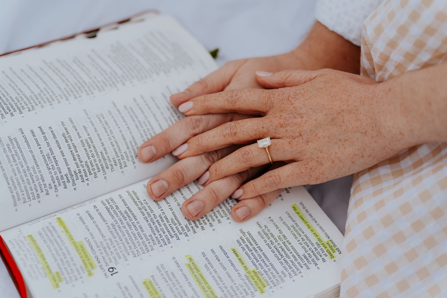 Hands resting on an open Bible with highlighted verses, wearing a gold ring, placed on a checkered cloth.