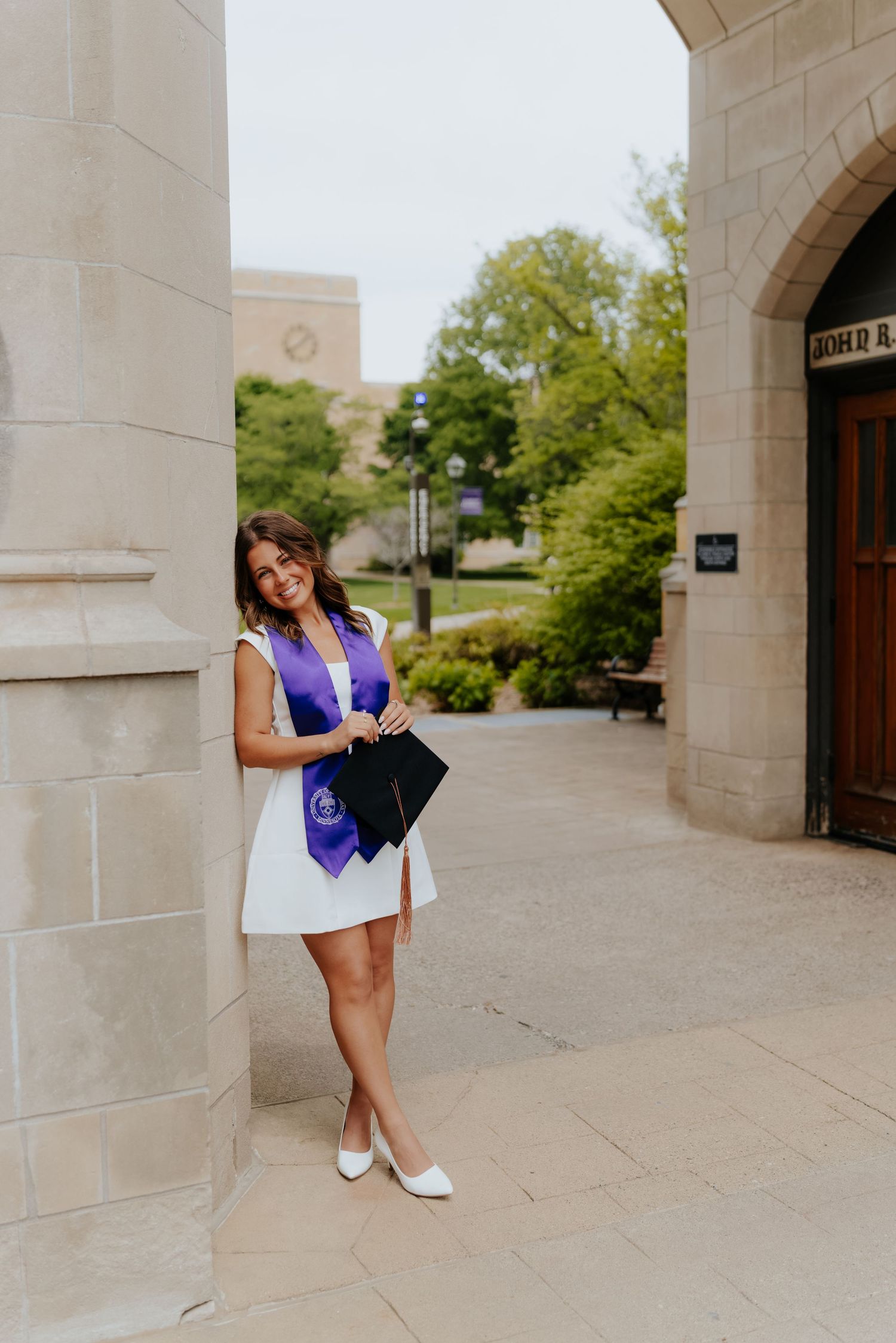 Young woman in white dress and purple graduation stole holds cap, posing by stone columns on college campus.