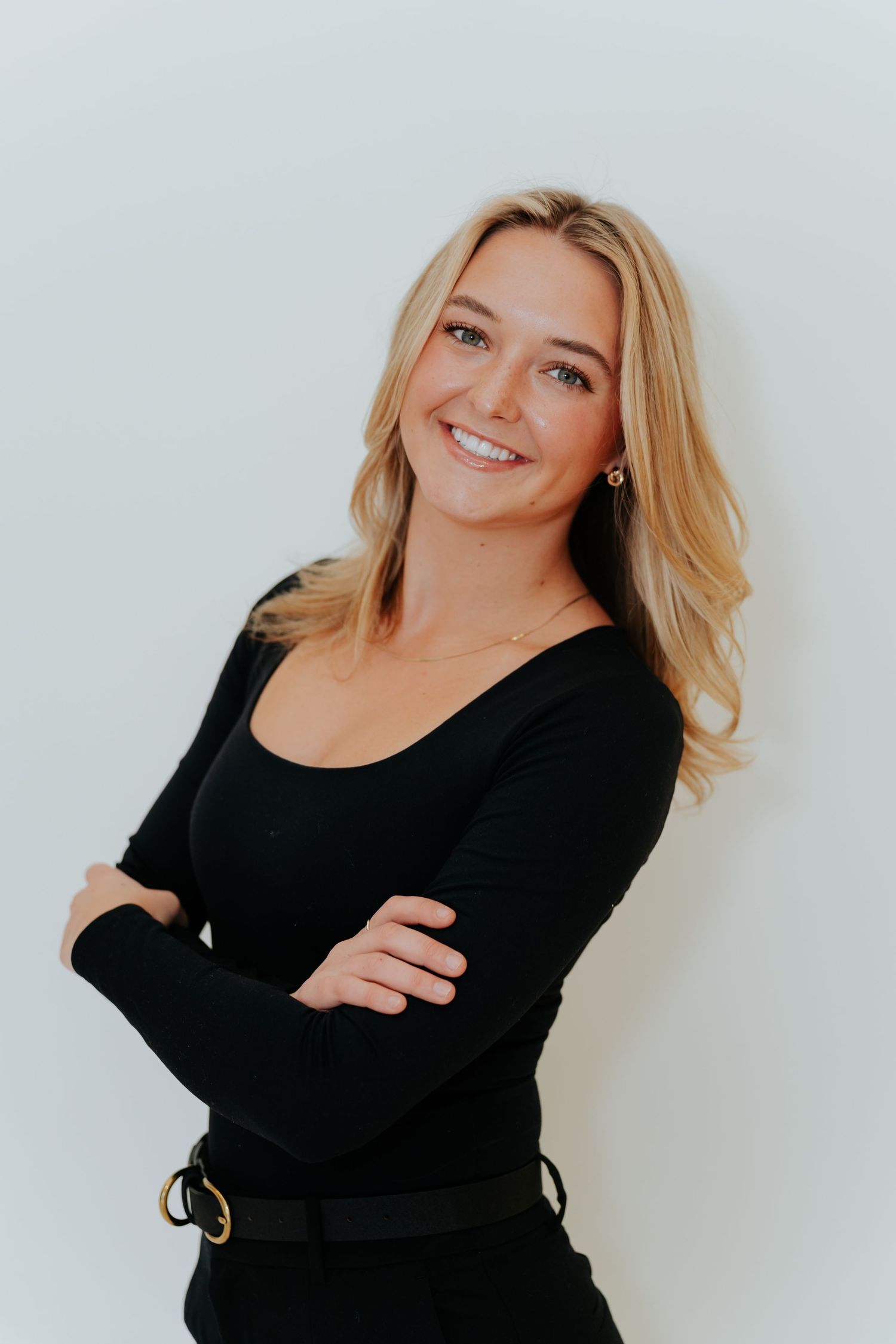 Smiling blonde woman in black long-sleeve top with arms crossed, posing confidently against a white background.