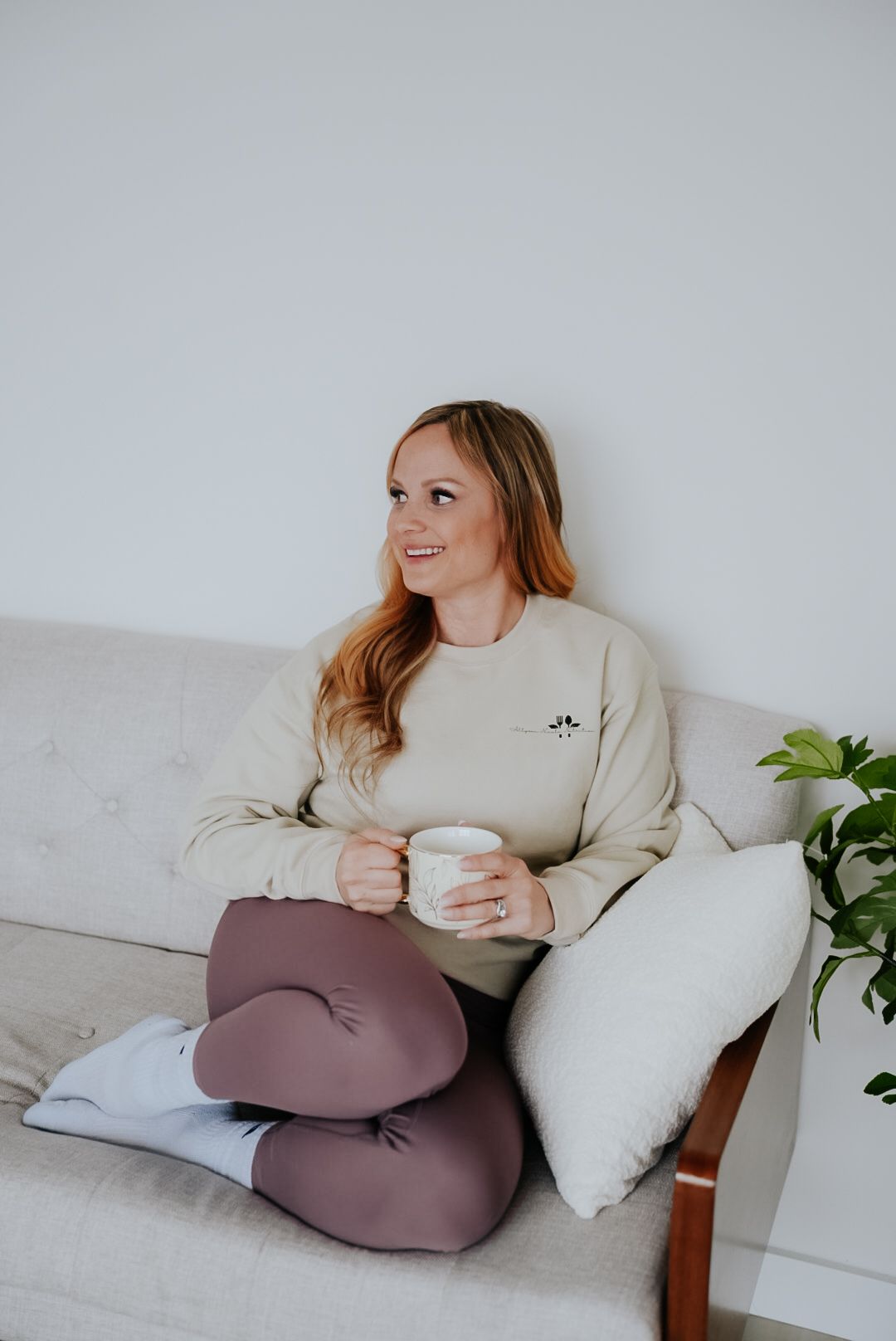 Woman in beige sweatshirt and mauve leggings sitting on couch holding white mug, smiling in bright living room.