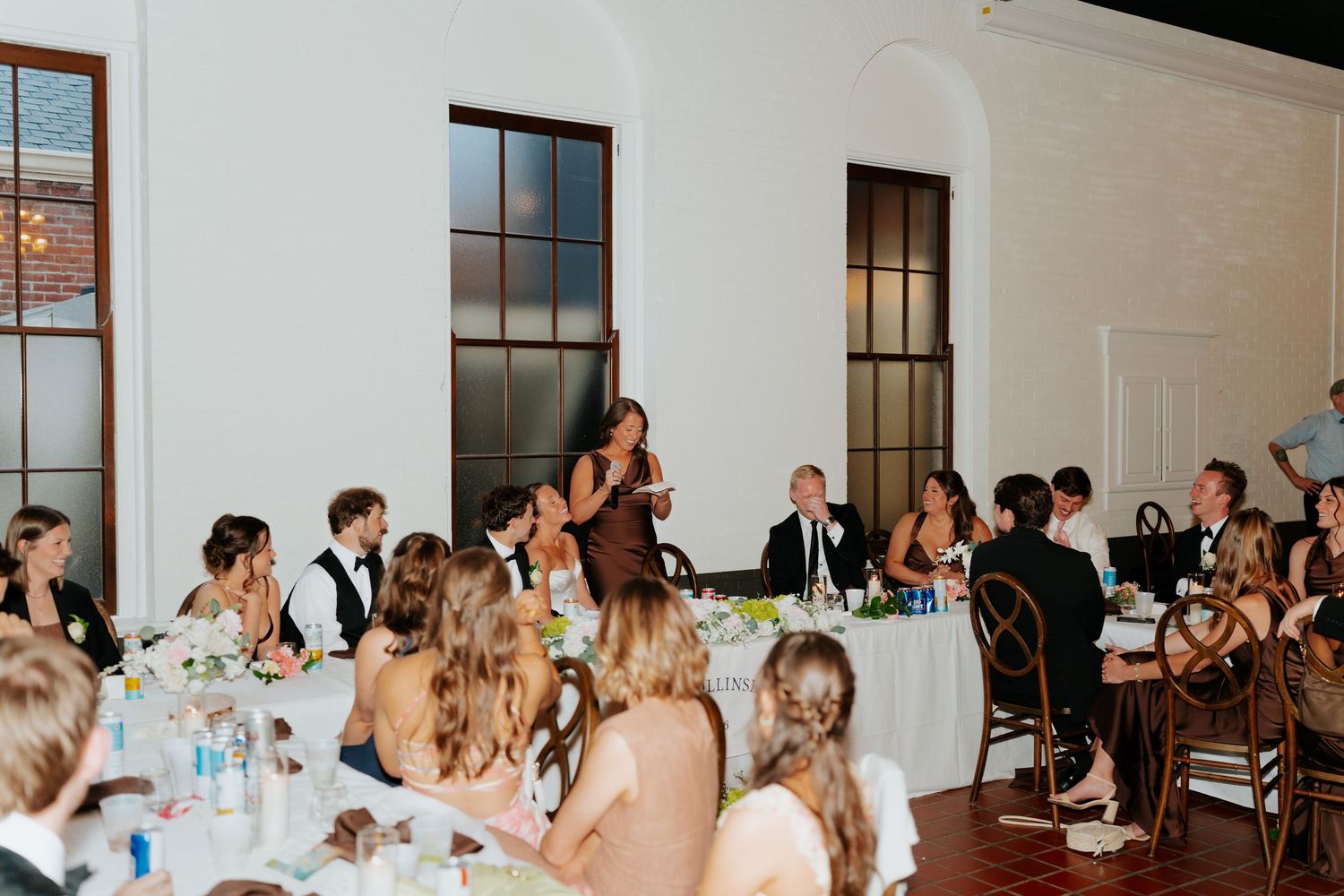 Wedding reception toast with guests seated at elegant tables in a white venue with large windows.